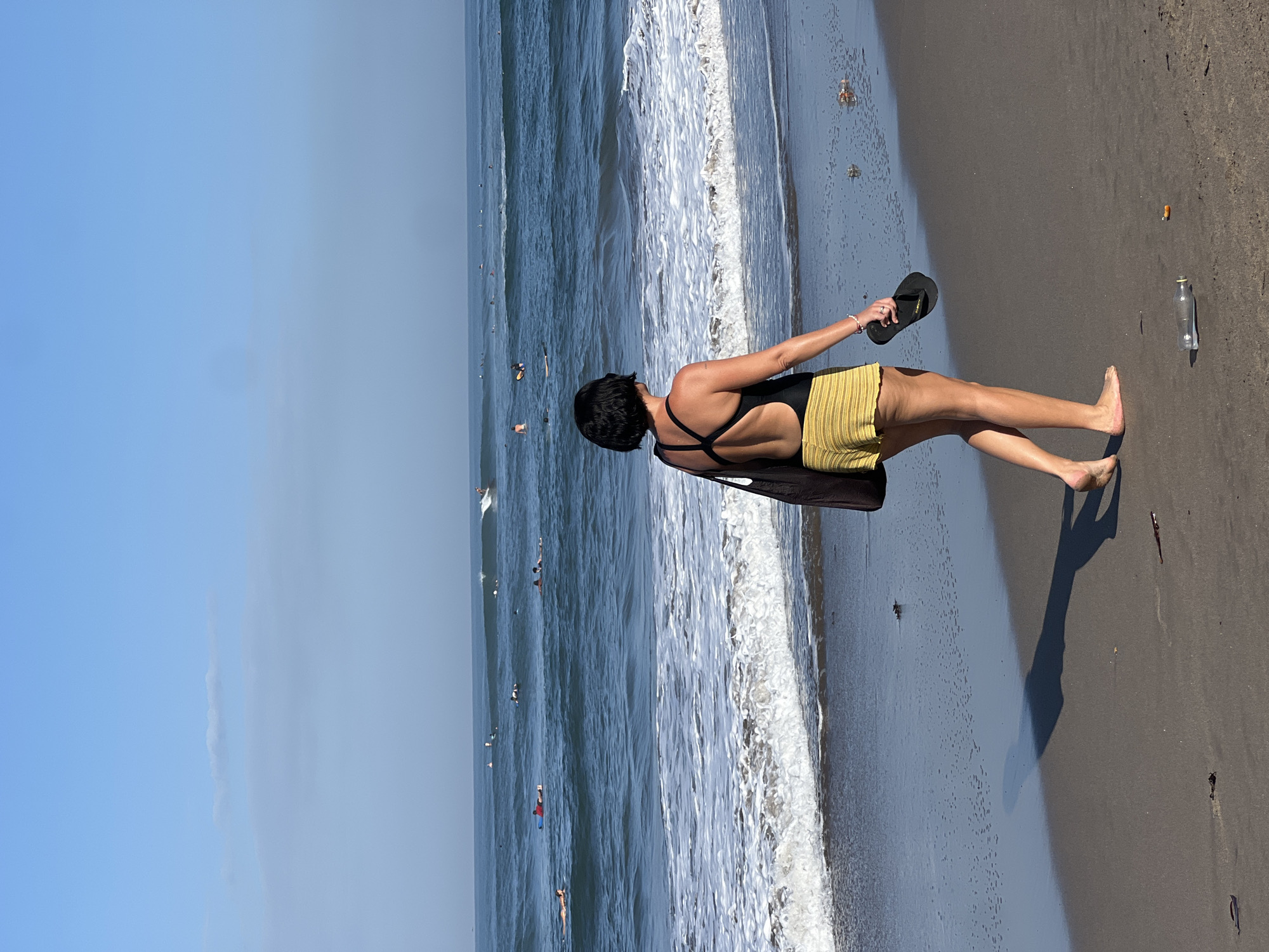 A woman with short black hair, wearing a black one-piece swimsuit and yellow striped shorts, walks along a wet sandy beach. She carries a dark tote bag on her left shoulder and a black flip-flop in her right hand. The ocean stretches out before her with numerous surfers in the water. The sky is a clear, light blue with a few wispy clouds. The scene is set on the beach in North Kuta, Indonesia, as indicated by the context.