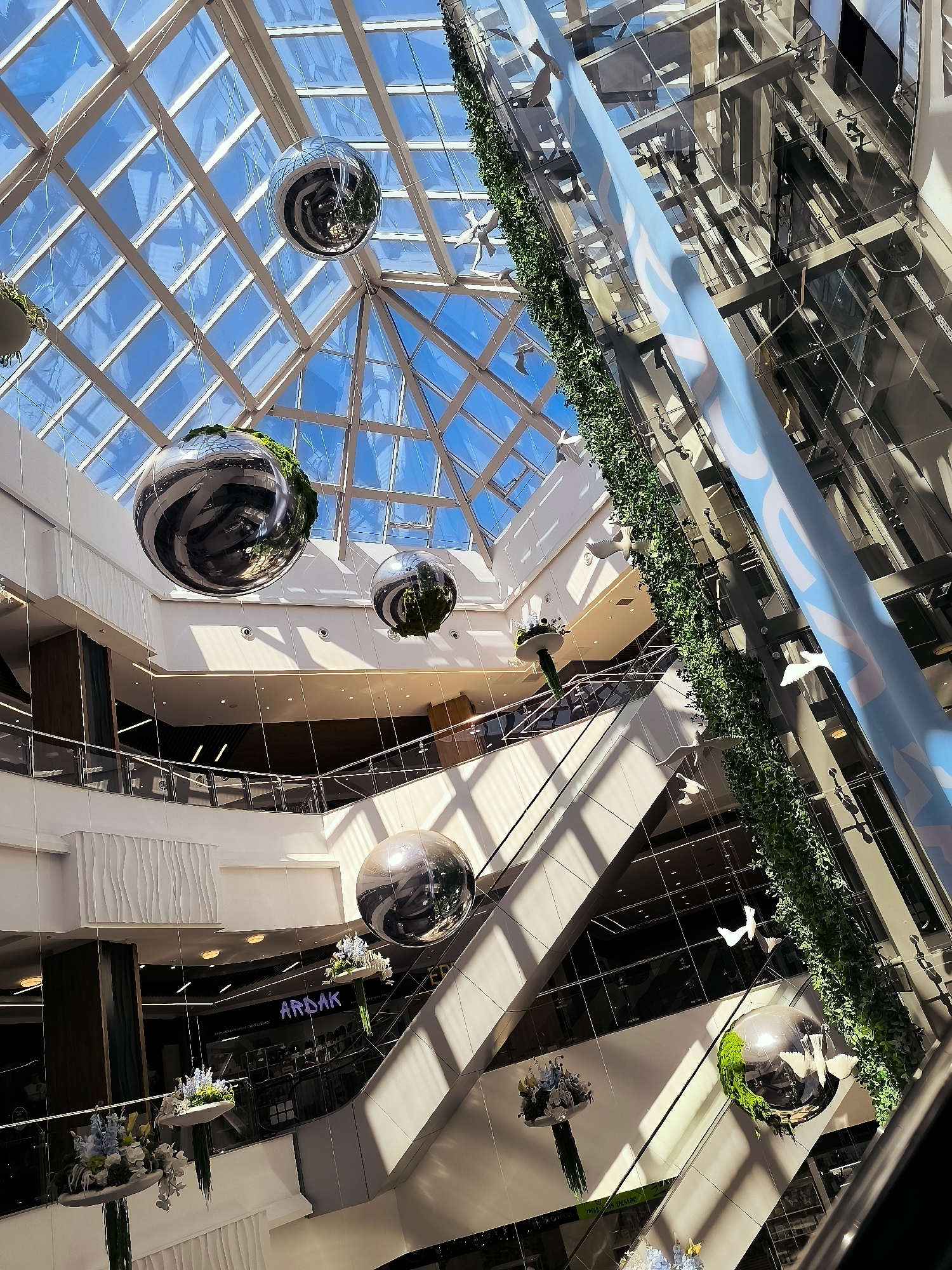 The image shows the interior of a modern shopping mall, likely in Qarağandı city administration, Kazakhstan. The atrium features a glass ceiling with a grid pattern, allowing sunlight to illuminate the multi-level structure. Large, reflective spherical decorations, some adorned with greenery, hang suspended from the ceiling by wires. A glass elevator shaft runs along one side of the atrium, with a decorative green wall and white bird figurines attached to it. Escalators and walkways are visible on different levels, connecting various sections of the mall. Signs with Cyrillic text are present on the lower levels, one of which reads "ARDAK". Floral arrangements hang from the ceiling on some levels. The overall scene suggests a bright, spacious, and contemporary commercial environment.