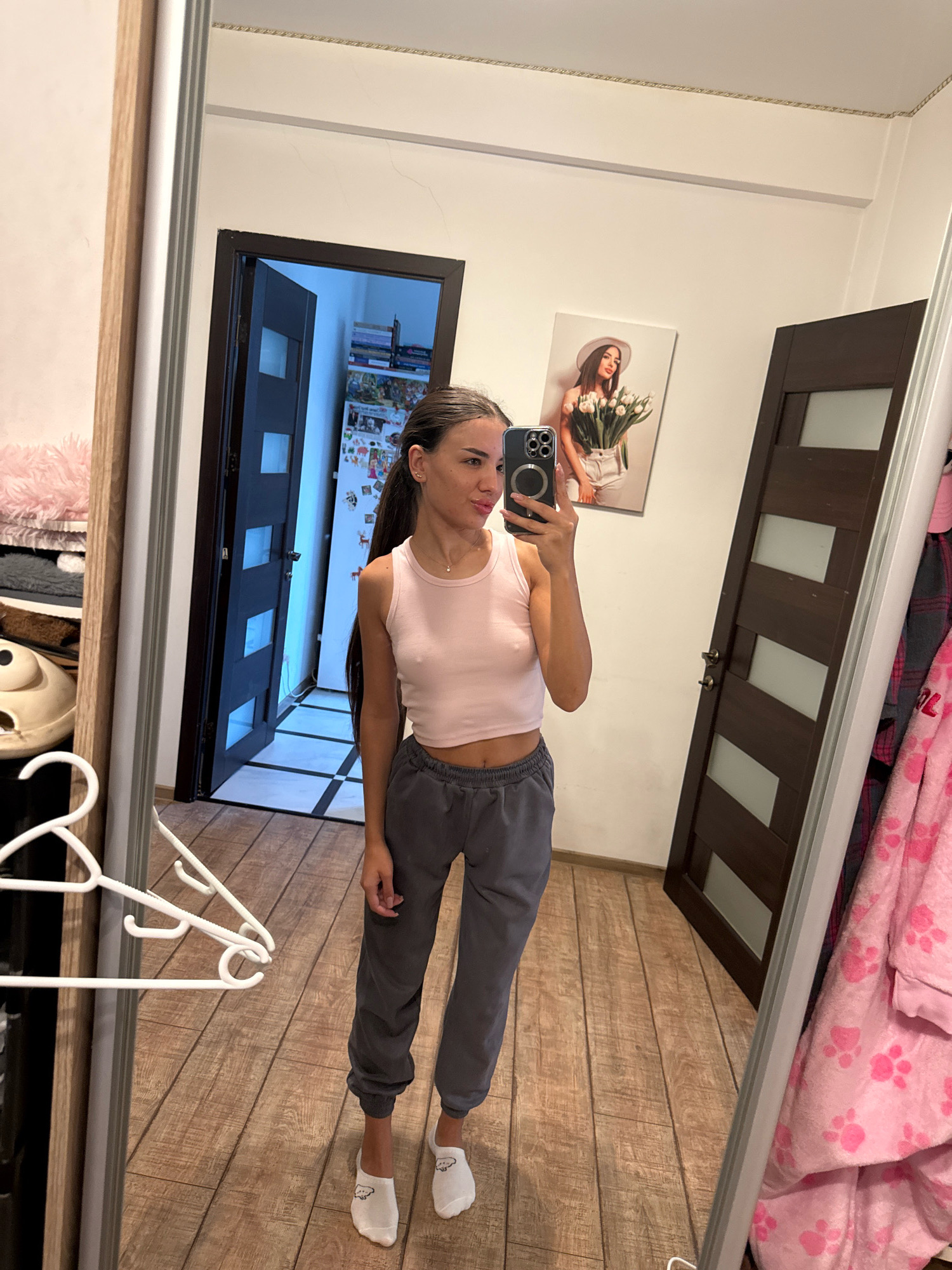 A young woman with long dark hair is taking a mirror selfie in what appears to be a home interior. She is wearing a light pink tank top and grey sweatpants, along with white socks with cartoon designs. The woman is holding a smartphone in her right hand, pointed towards the mirror. In the background, a doorway with a checkered floor is visible, along with two dark wooden doors. There is also a framed picture of a woman holding tulips on the wall and a large mirror to the right. The flooring is made of wooden planks. There is no visual information to suggest the location is in Chișinău, Moldova.