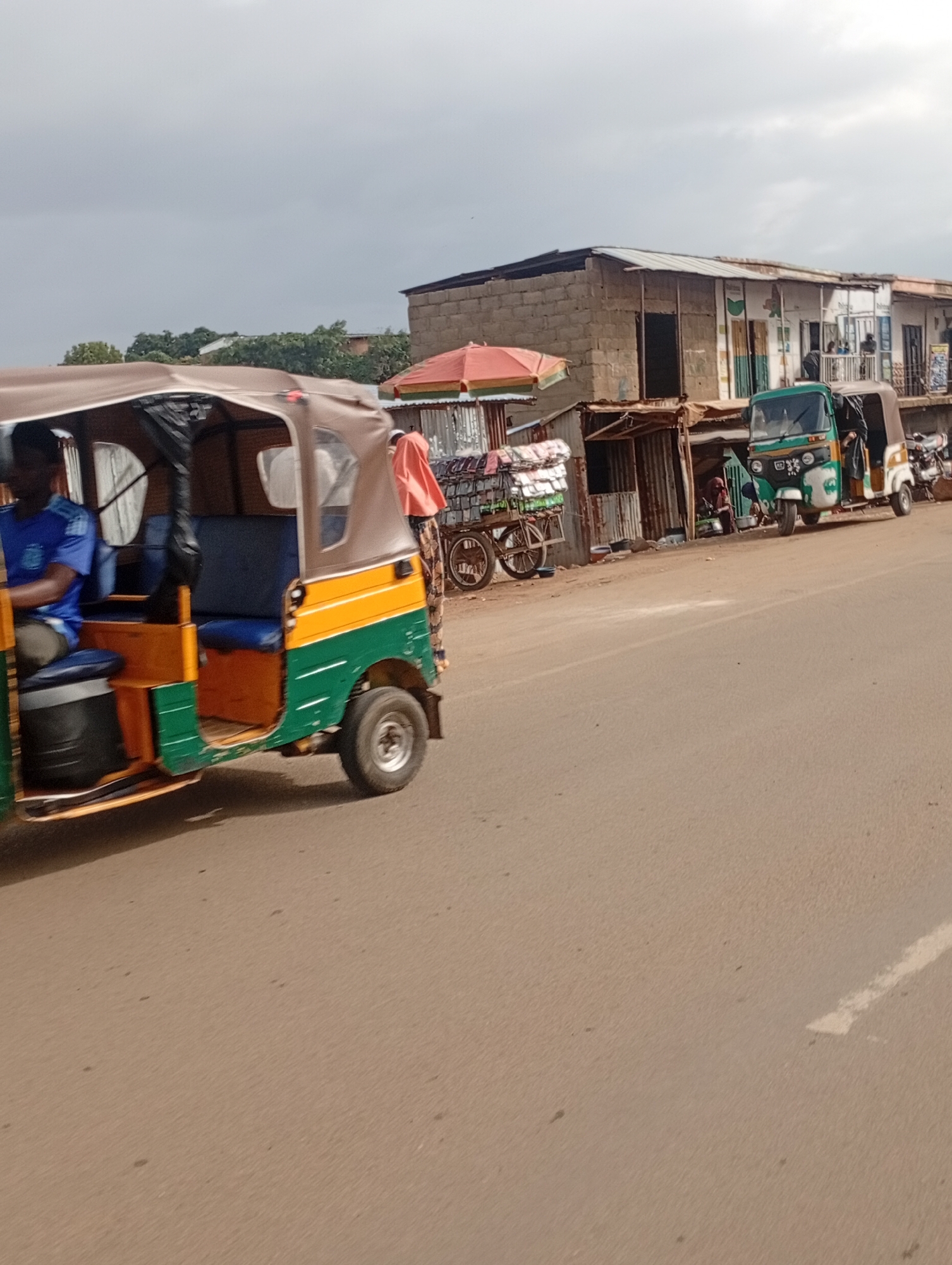 This media file shows a street scene in Rafin Par, Nigeria. The foreground is dominated by a colorful auto-rickshaw, a three-wheeled vehicle. It is primarily green and yellow, with a brown canopy. A person wearing a blue shirt is visible in the driver's seat. The auto-rickshaw appears to be moving or parked on a paved road.
In the background, there are several small shops and buildings lined up along the roadside. These structures have corrugated metal roofs and appear to be constructed from brick and other materials. Some of these shops have open fronts, revealing goods and people inside. One shop has a red and yellow umbrella set up outside.
There are other vehicles visible, including another auto-rickshaw that is green and white, parked further down the road. A person wearing a pink headscarf and patterned skirt is standing near this vehicle. A motorcycle is also parked in front of one of the buildings. In front of one of the shops, there is a cart loaded with stacked items, possibly goods for sale.
The sky is overcast, suggesting cloudy weather. The lighting indicates it could be daytime, perhaps late morning or afternoon. The scene suggests a bustling local marketplace or a roadside commercial area, typical of many towns in Nigeria. There is no visible text in the image. The overall impression is one of everyday life and commerce in a Nigerian community.