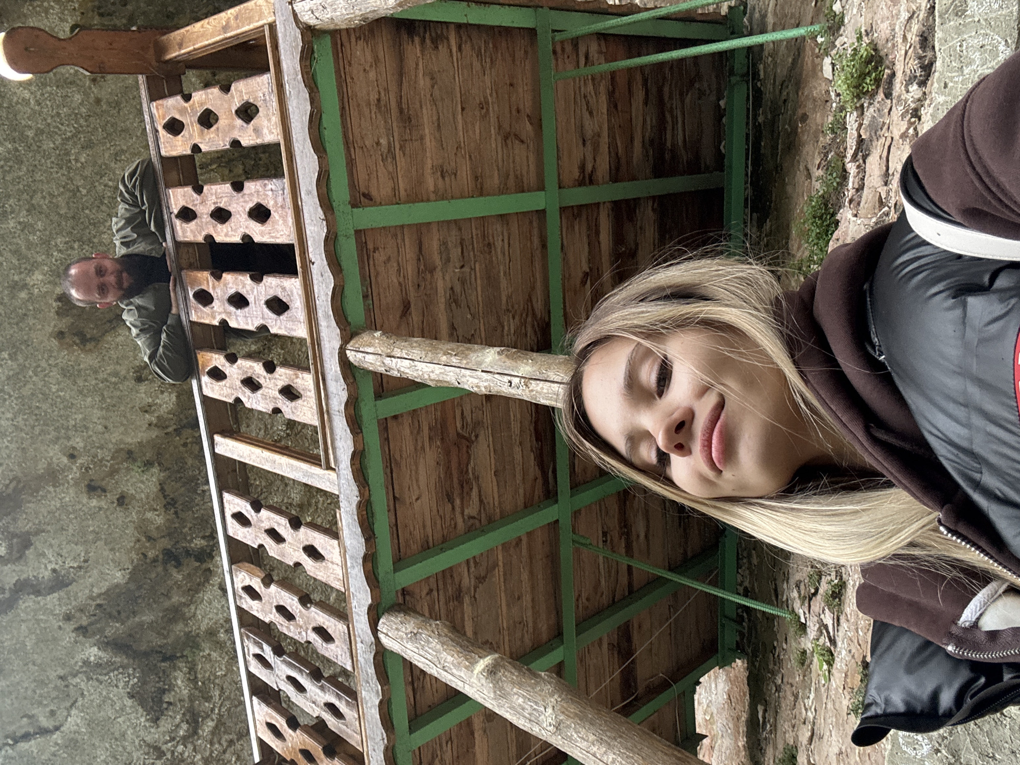 The image shows a young woman with blonde hair in the foreground, wearing a brown hoodie and a black jacket. She is smiling slightly. Behind her is a wooden structure with a green frame and patterned railings. In the background, through an opening in the structure, a man with a beard is visible. The structure appears to be located in a natural environment, possibly a rocky or mountainous area, with some greenery visible on the right. The photograph was taken in Tsabal, Abkhazia.