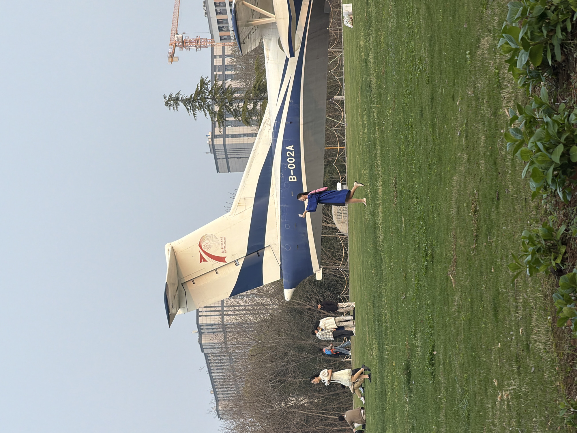 The image shows a static display of an aircraft in Jiangning District, China. The plane's tail section is prominently visible with the registration number B-002A. A woman dressed in a dark blue robe is standing beside the fuselage. In the background, there are other people on the grass, some standing and others sitting. There are buildings behind the people, as well as a construction crane. There is a green lawn in the foreground with shrubbery along the right edge.