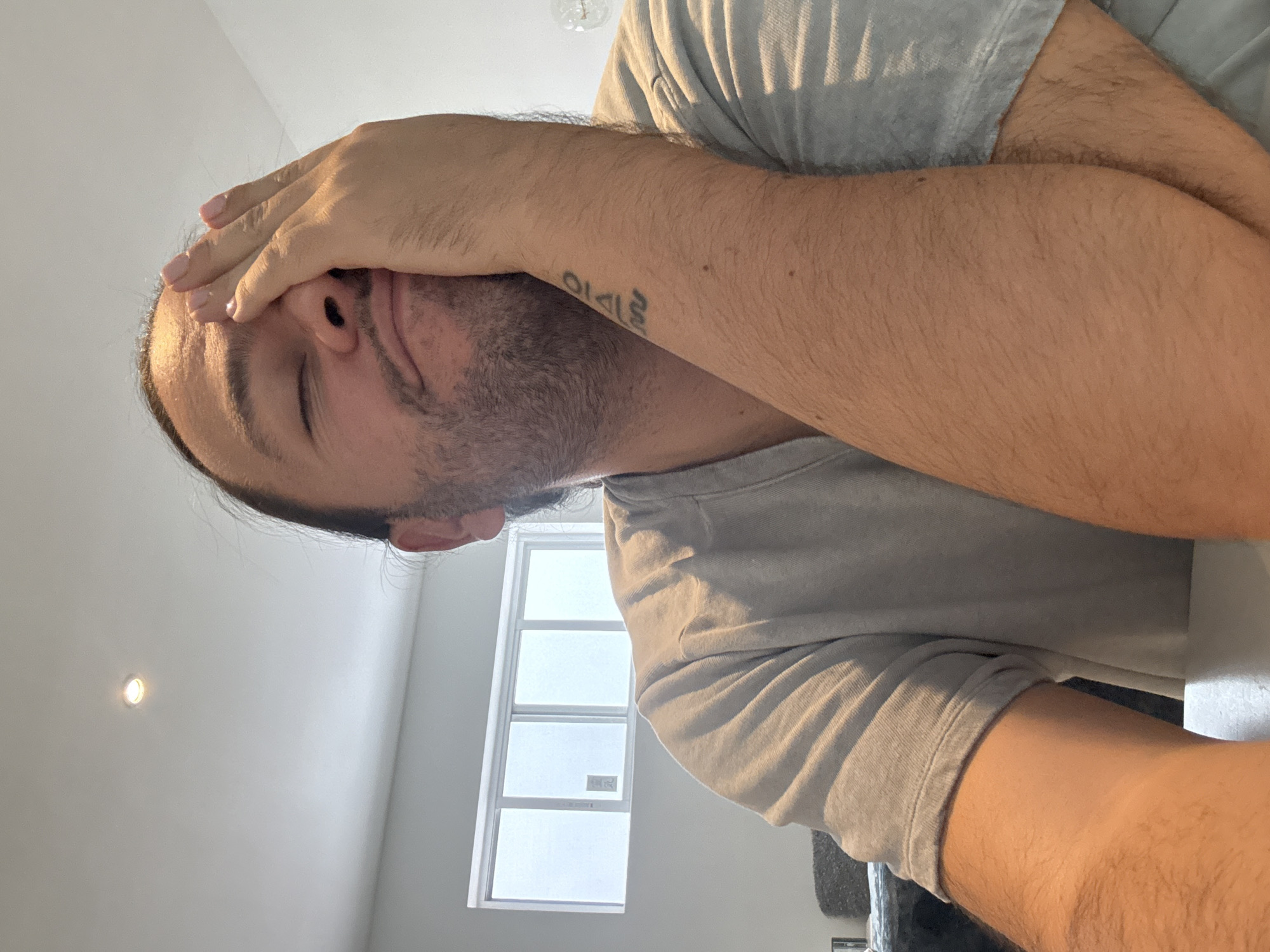 A man covers his face with his hand in a brightly lit room. He has a beard and a tattoo on his arm. A window is visible in the background. There is no evidence to suggest the location is Puebla, Mexico.