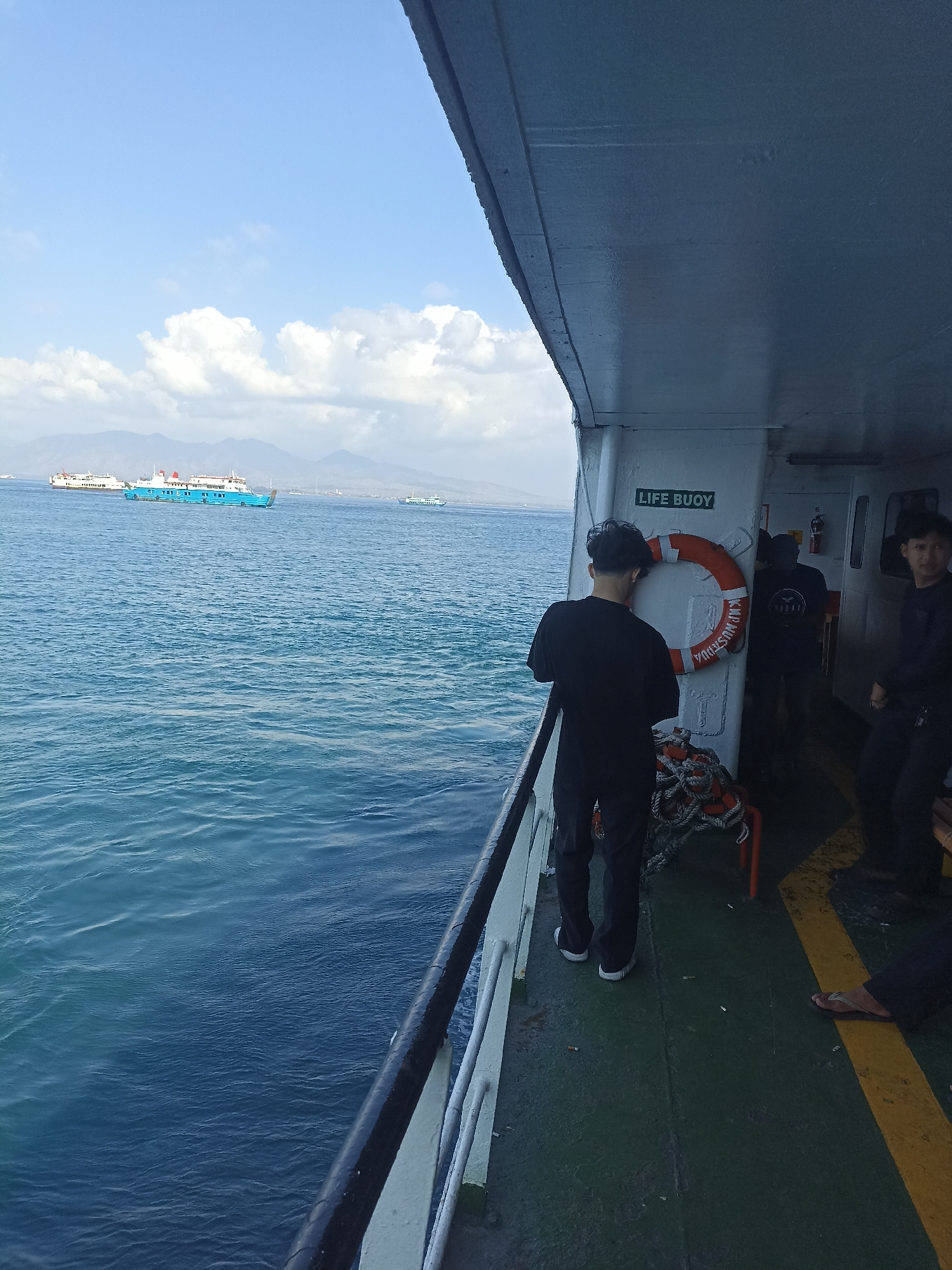 On a ferry, likely en route to or from Ketapang, Indonesia, three young men stand on the deck. The man closest to the camera, wearing a black t-shirt and pants, faces away from the viewer, looking out at the ocean. He is positioned on the port side of the ferry, with a railing separating him from the water. To his right, another man in a black t-shirt and dark pants stands further back, also looking towards the water. A third man, also in dark clothing, stands on the far right of the frame, his gaze directed forward. A bright orange life buoy, labeled "LIFE BUOY" in green lettering, hangs on a white bulkhead behind the first man. The ferry deck is a greenish-gray color with a yellow stripe marking the edge. Scattered cigarette butts are visible on the deck. The ocean is a deep blue with gentle waves, and in the distance, several other ferries and landmasses with mountains are visible under a bright blue sky with fluffy white clouds.