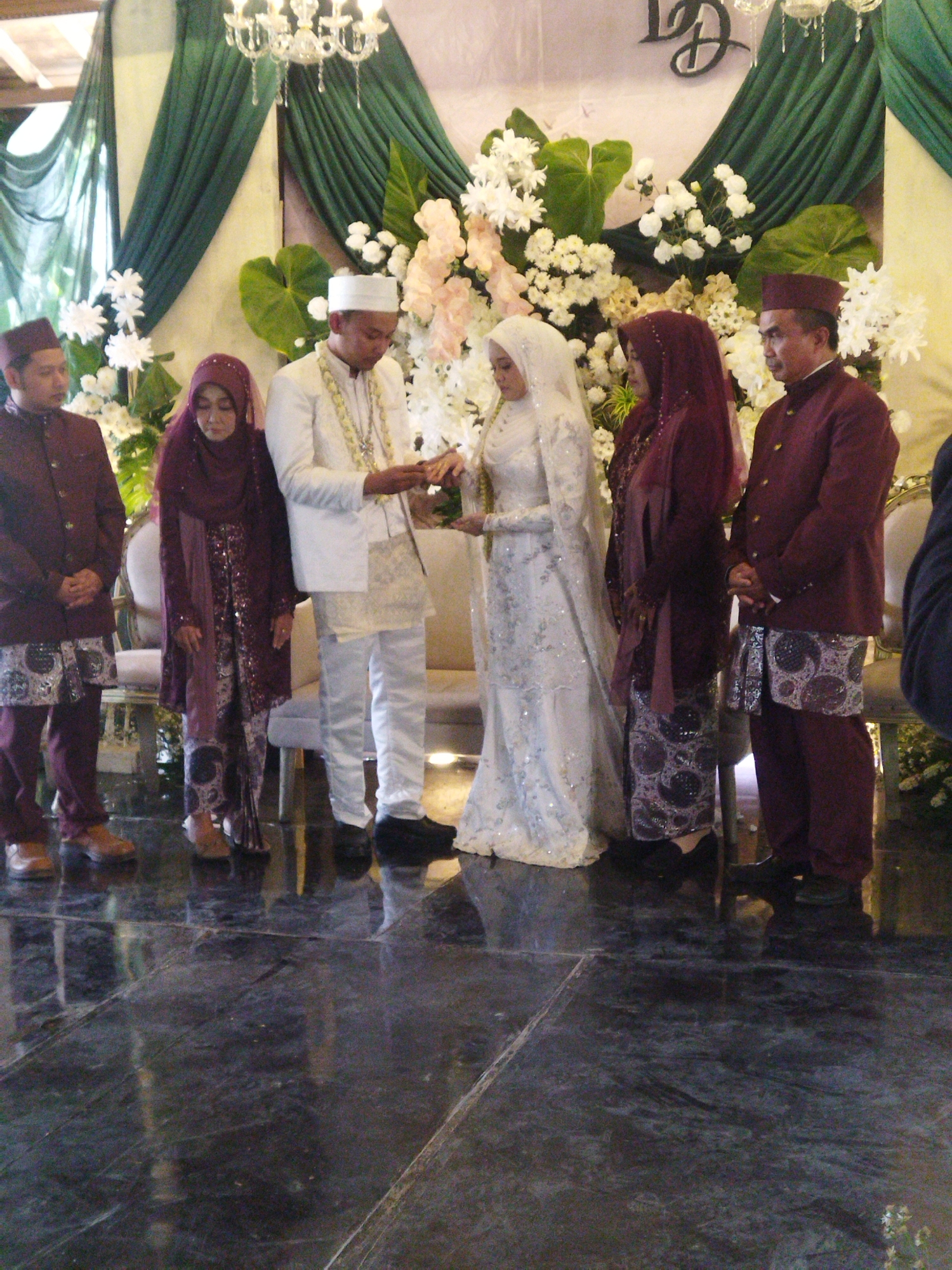A wedding ceremony is taking place in what appears to be an indoor venue, possibly a banquet hall or event space, adorned with dark green drapes and white floral arrangements. The event is being held in Semarang, Indonesia. A wedding couple, dressed in traditional attire, stands at the center. The groom wears a white suit with a traditional peci hat, while the bride is in a white gown and hijab, adorned with intricate beadwork. They are exchanging rings or performing a ritualistic hand-holding, suggesting the exchange of vows. Flanking them are four other individuals, two on each side. On the groom's right are two men in maroon traditional wedding outfits, consisting of jackets and pants with ornate sarong-like fabrics around their waists. On the bride's left are two women, one in a maroon hijab and matching dress, and another in a similar outfit. The flooring is a dark, reflective surface, possibly marble or polished stone, showing reflections of the people and the surroundings. The overall atmosphere suggests a formal and culturally significant event.