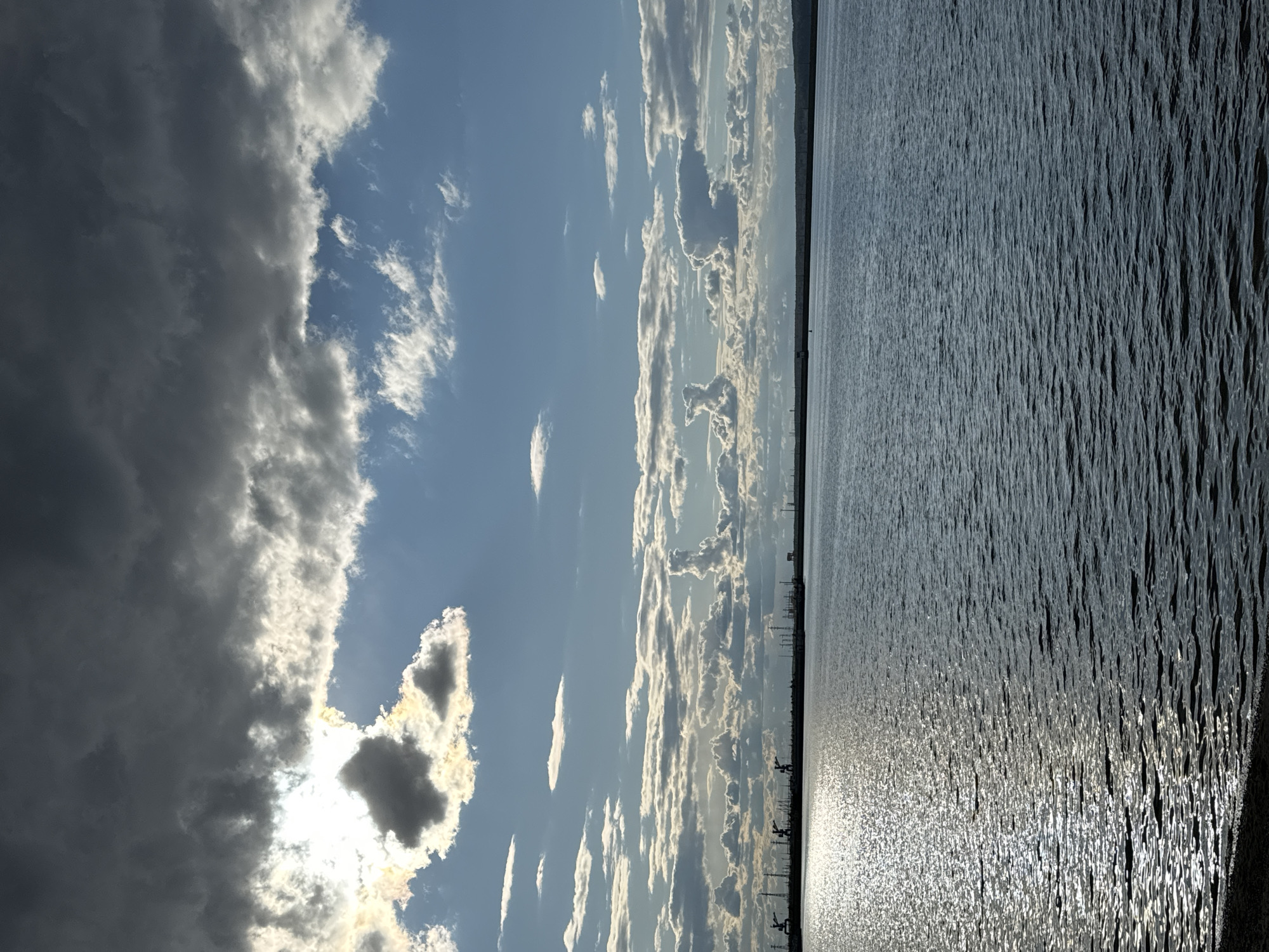 The image displays a vast waterscape under a dramatic, cloudy sky. A large body of water, likely a river or reservoir, occupies the lower half of the frame, its surface a textured expanse of dark grey-blue with visible ripples and small waves. A brilliant strip of sunlight shimmers intensely on the water towards the right, indicating the sun's position.
The sky above is filled with varied cloud formations. On the left, heavy, dark cumulus clouds dominate, partially obscuring the sun, which casts a bright, luminous halo from behind them. Moving towards the right, the sky becomes a lighter blue, interspersed with numerous smaller, whiter, and brighter cumulus clouds that catch the sunlight.
A clear, straight horizon line separates the water from the sky. Along this distant line, faint silhouettes of industrial structures, possibly cranes or pylons, are visible, suggesting the proximity of an urban or industrial area. The overall lighting, with the low-lying sun and the interplay of bright reflections and dark clouds, suggests either late afternoon or early morning. There are no people or discernible activities taking place, and no visible text. The scene portrays a wide, open landscape consistent with a large body of water near Chaikovsky, Russia.