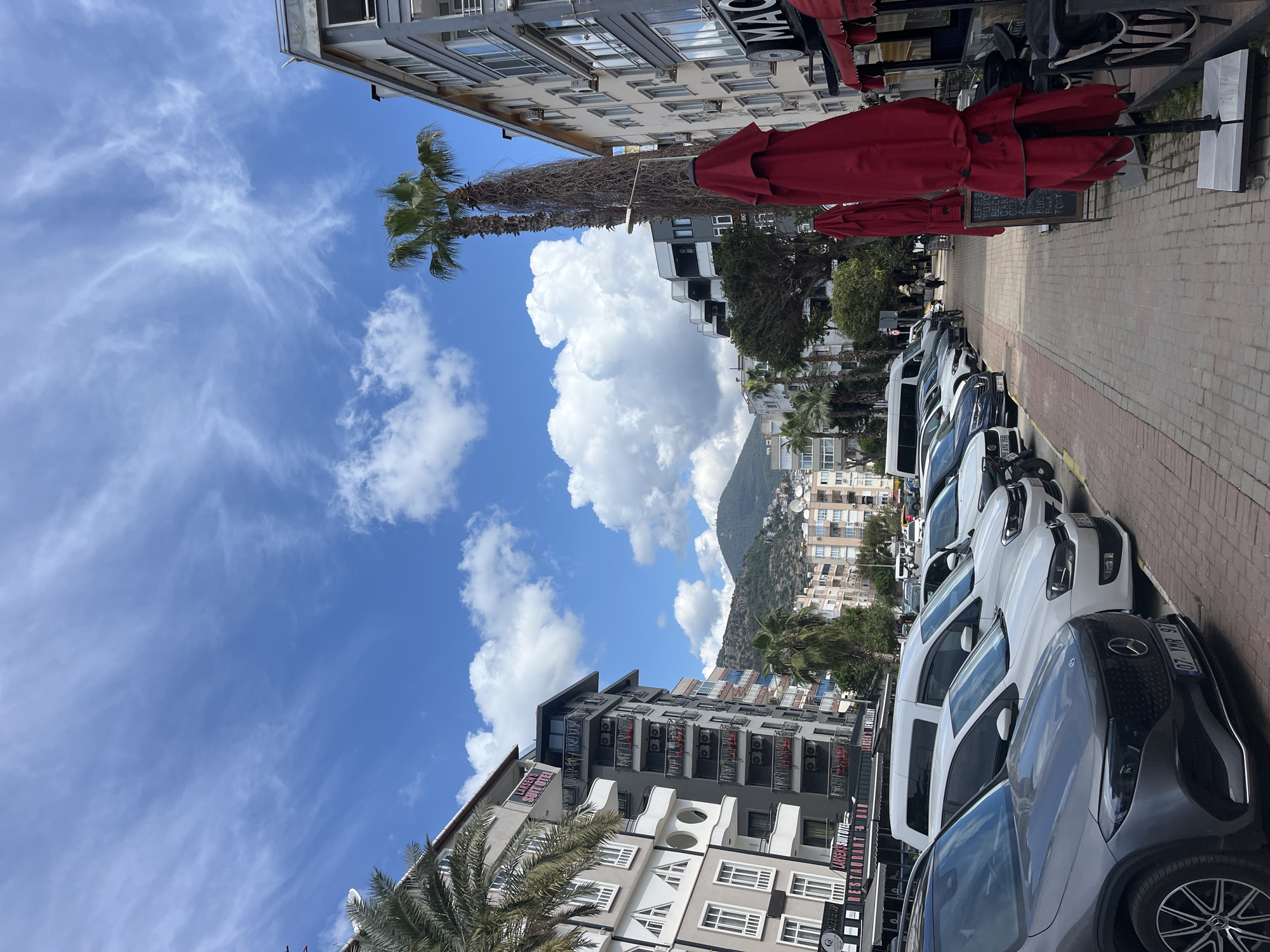 The image shows a street in Alanya, Turkey, lined with parked cars, buildings, and a palm tree. The sky is blue with large white clouds. A mountain is visible in the background. A restaurant with red umbrellas is on the right side of the street. Buildings of different heights and colors (gray, white, with dark balconies) are located on both sides of the street. The license plates of some cars are visible.