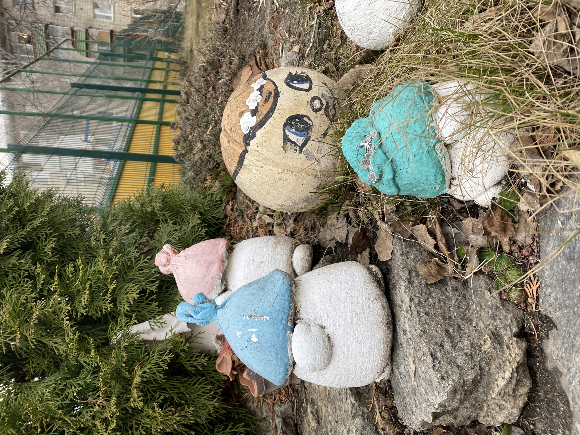 The photograph shows a small outdoor garden or landscaping display.
Several painted rocks are arranged among vegetation and fallen leaves. A round rock painted with a crude face is prominent. Other rocks are painted white with colored tops, including pink, blue, and turquoise. A green coniferous bush is visible on the left side of the frame.
In the background, a fenced structure with yellow panels is partially visible. A multi-story building stands in the background as well.
The image was taken in Yekaterinburg, Russia.