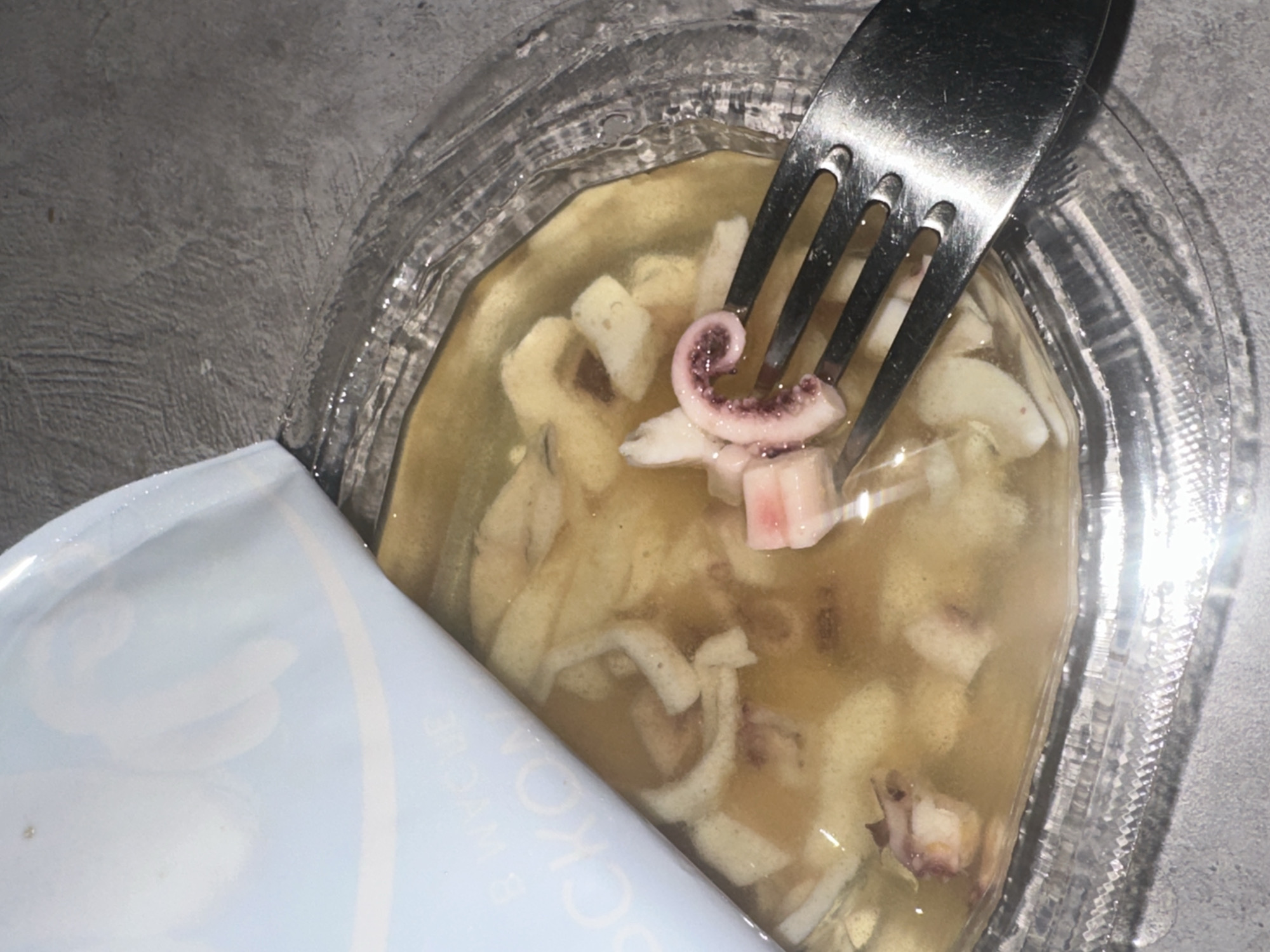 An open container of what appears to be pickled squid or octopus sits on a gray surface. The container is clear plastic with a white and light blue lid partially peeled back. Inside, the seafood sits in a yellowish brine. A silver fork is inserted into the container, holding a piece of squid. The environment is bland and lacks any distinguishing features that would provide clues to a specific location in Chaikovsky, Russia or elsewhere.