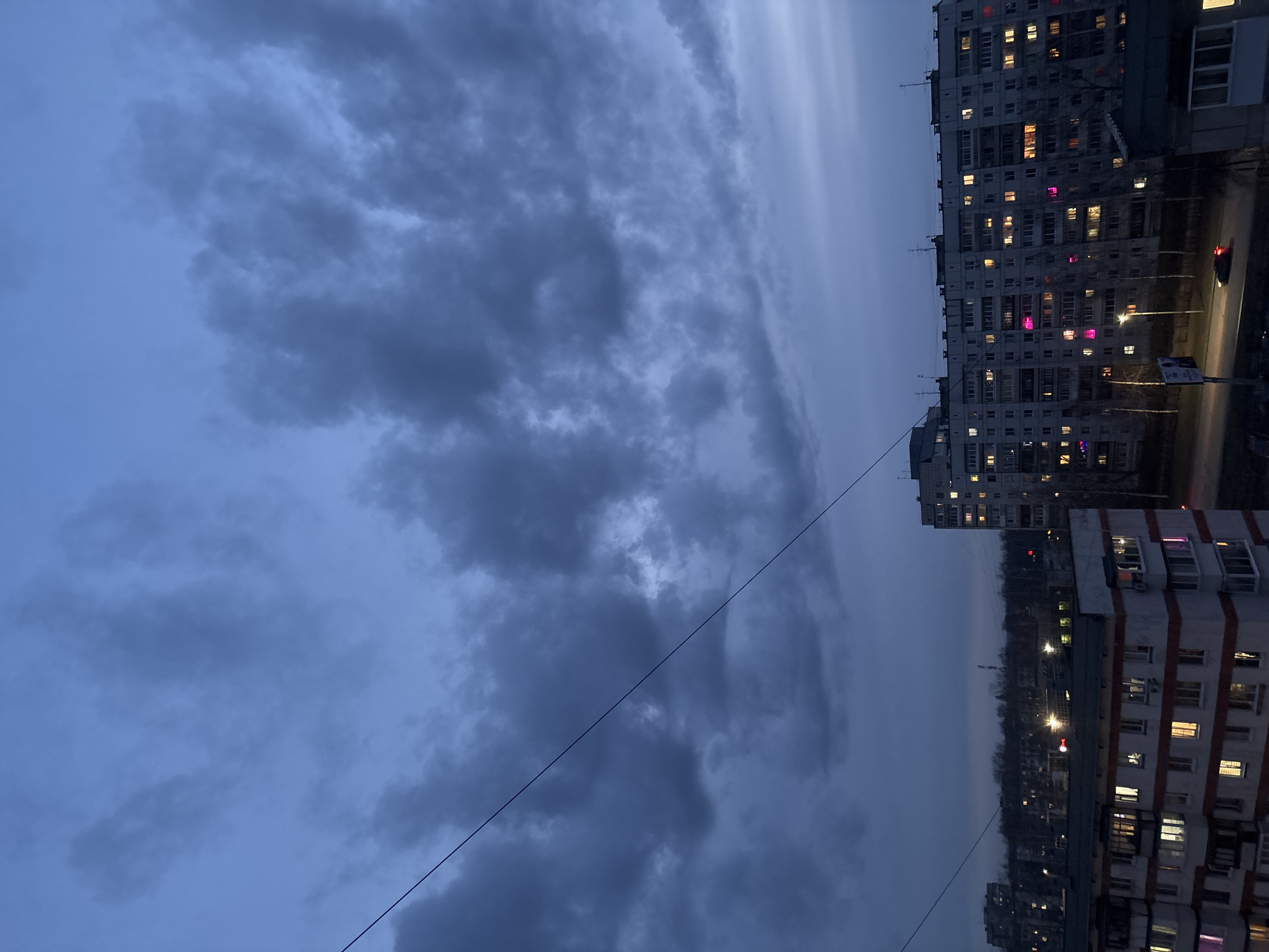 The image shows a nighttime view in the city of Chaikovsky, Russia. The sky is overcast with dark, textured clouds. Power lines are visible against the sky. In the background, there are several multi-story apartment buildings with many illuminated windows. Streetlights illuminate the road in front of the buildings. The overall scene conveys a sense of urban twilight or early evening.