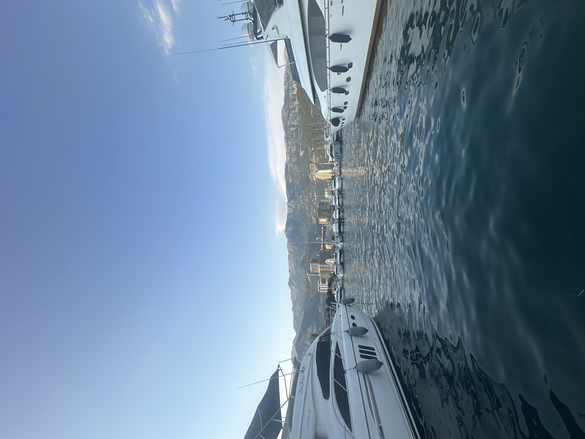 This is a wide-angle, full-shot photograph of a marina in Budva, Montenegro, taken from the water. The scene is dominated by two large white yachts, one on the left and one on the right, framing the view. Both yachts are moored and appear to be unoccupied. The water in the foreground is a deep blue-green and shows gentle ripples. In the background, a city skyline with modern buildings is visible, nestled at the base of a large mountain range. The sky is clear and light blue, with a few wispy clouds on the right side. The overall impression is one of a serene and picturesque coastal environment, characteristic of Budva's harbor area, possibly near the Old Town or the modern marina.