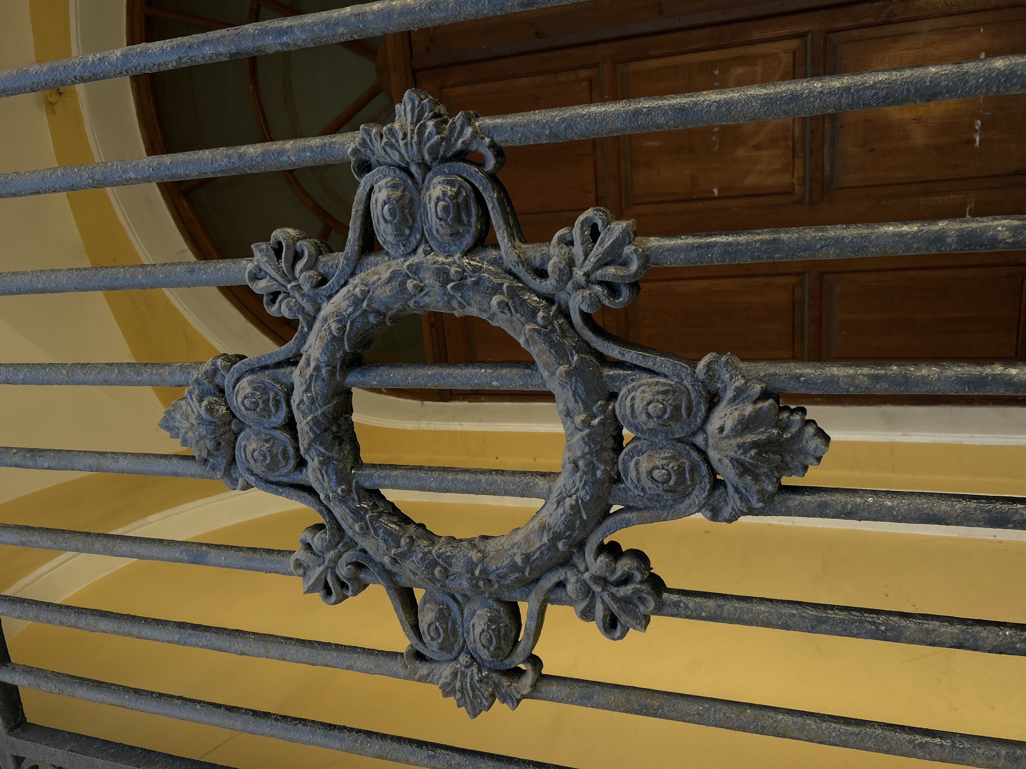 The image displays a close-up view of an ornate, dark metal grate or railing. At its center is a decorative medallion, likely cast iron, fashioned into a circular, wreath-like shape adorned with symmetrical floral and foliate motifs. The metal appears weathered and textured, suggesting age and intricate craftsmanship.
The grate itself is composed of multiple horizontal metal bars. Behind this ironwork, a soft, warm yellow architectural surface, possibly a curved wall or arched ceiling, provides a contrasting background. Further in the distance, through the gaps in the grate, a section of wooden paneling with distinct rectangular patterns is visible. To the upper left, a segment of a round window with wooden frames adds depth to the background.
Soft, even lighting illuminates the textures of the metal and wood, suggesting an interior or sheltered exterior space during daytime. The scene evokes a sense of historical architecture and artistic detail, characteristic of its location in Saint Petersburg, Russia. No people or visible text are present in the photograph.