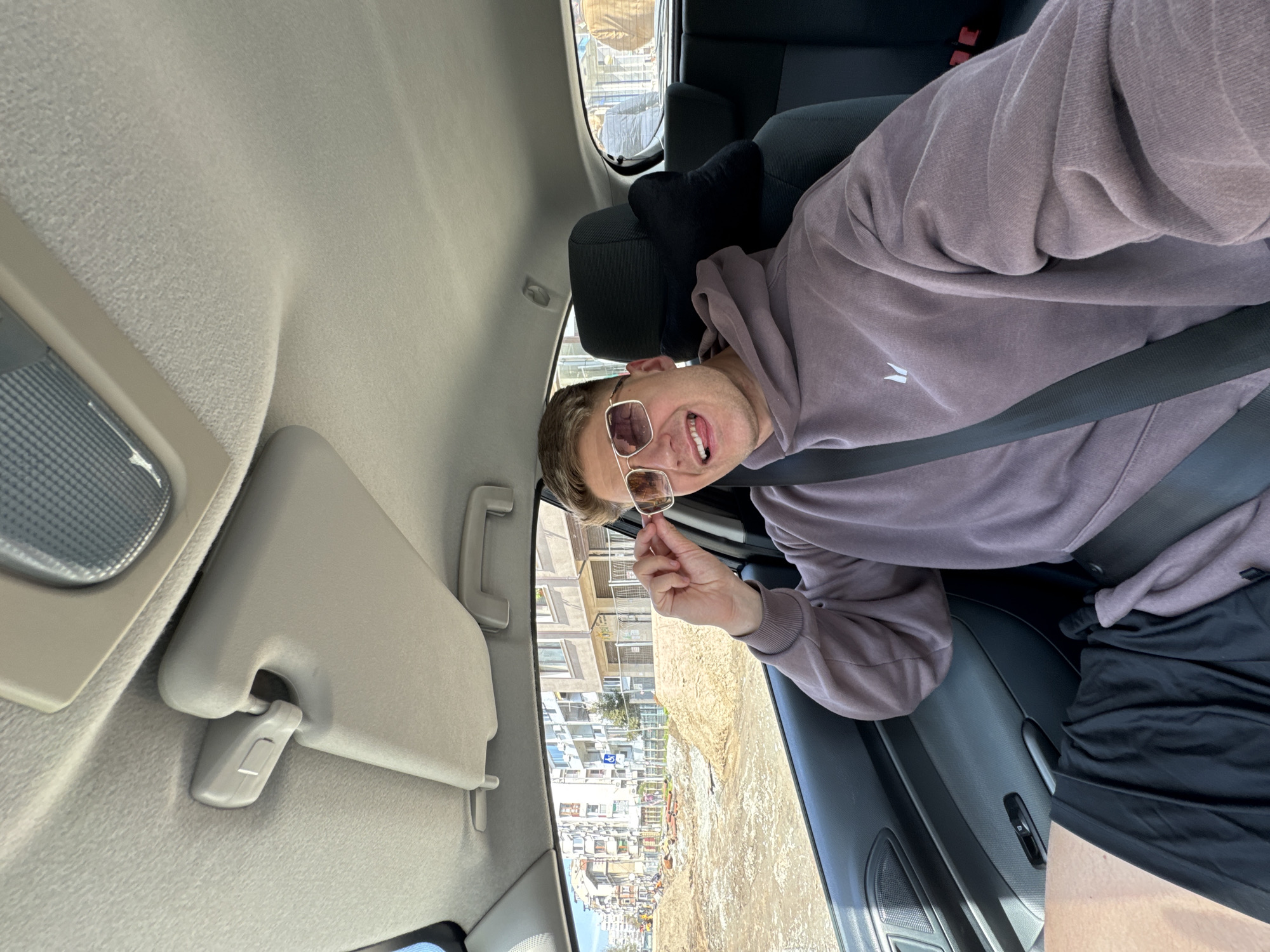 A person is sitting in the driver's seat of a car. They are wearing a mauve-colored hoodie and black athletic pants. They have on sunglasses and are holding them by the frame in their right hand. They are looking at the camera. A seatbelt is fastened. The car interior is gray. The sun visor is down. Through the car window, a street in Burgas, Bulgaria is visible. Construction work is ongoing. Buildings line the street.