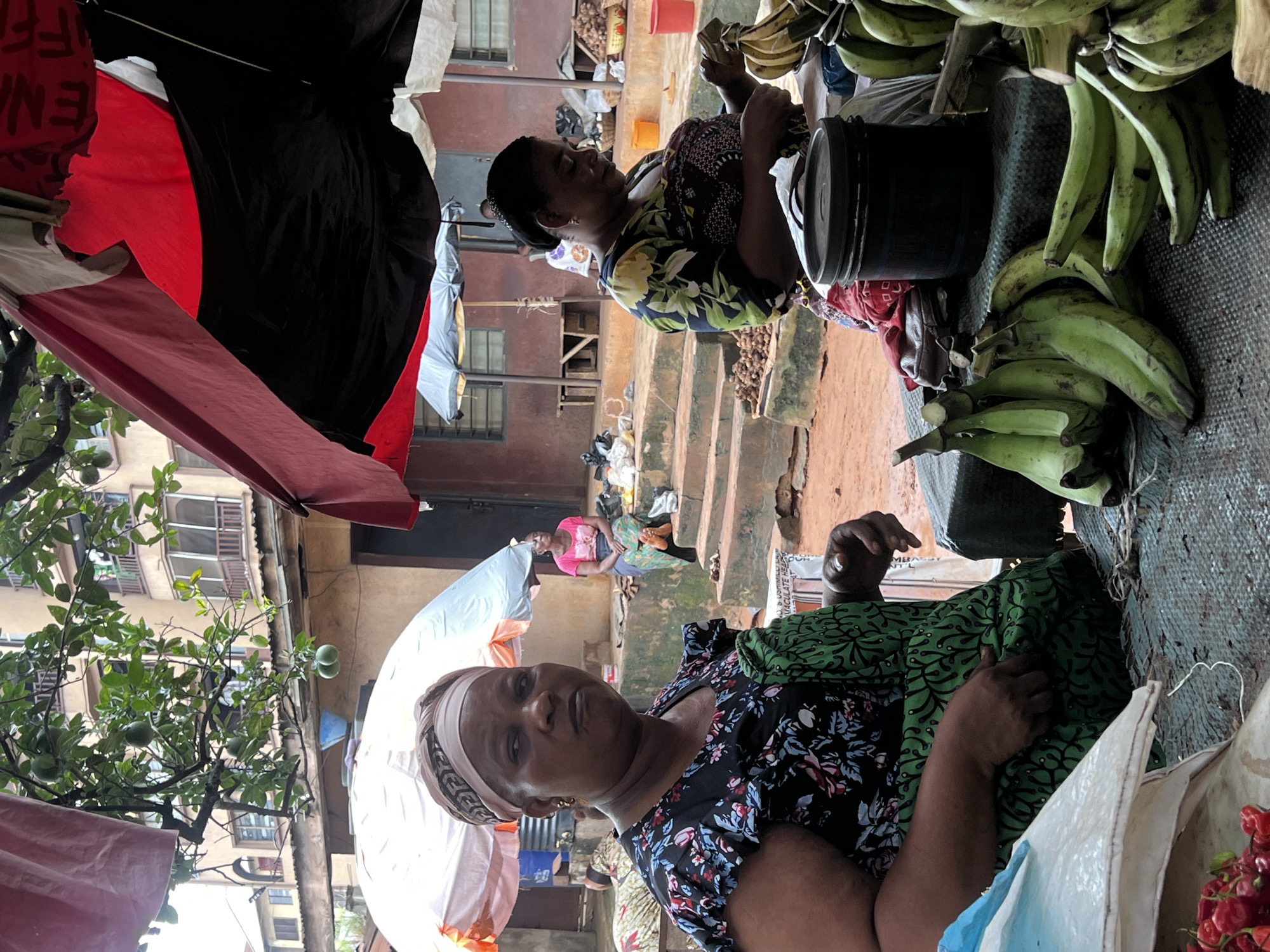 The image depicts a bustling outdoor market scene in Nkpor, Nigeria. In the foreground, a woman with a headscarf and a floral-patterned dress is looking directly at the camera, her left hand resting on a green apron. To her right, another woman, also wearing a headscarf, is focused on preparing a bunch of green plantains. Behind them, a person in a pink shirt is seated on steps, and further in the background, a building with multiple balconies is visible. The market is covered by awnings, including a red and black one partially obscuring the top of the frame. The ground appears to be dirt, and various goods, including more plantains, peppers, and baskets of produce, are displayed.