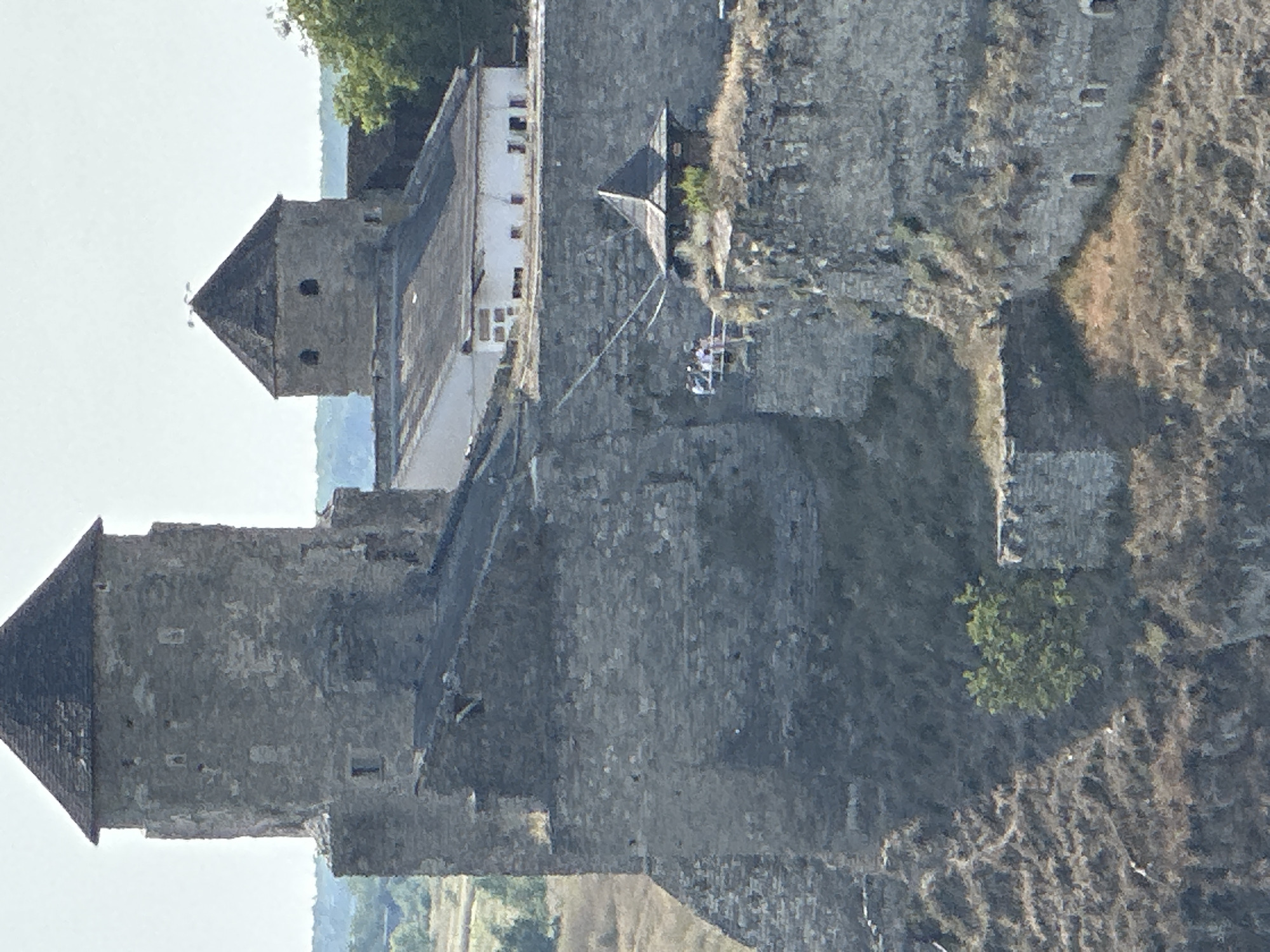 This is a medium-range, eye-level, exterior shot of the Kamianets-Podilskyi Castle in Kamianets-Podilskyi, Ukraine. The castle is a large, stone structure situated on a rocky outcrop. The main focus is on two towers; one large, cylindrical tower with a conical roof dominates the left side of the frame, while a smaller, octagonal tower with a conical roof is visible to the right, behind a building with a white facade and a dark roof. In the foreground, a section of the castle wall is visible, with a small, dark, hexagonal structure with a pointed roof on its upper surface. Several people, appearing to be tourists, are standing on a platform next to this structure. The surrounding landscape is dry and scrubby, with some sparse vegetation and trees visible in the background. The sky is overcast and a pale blue-grey.