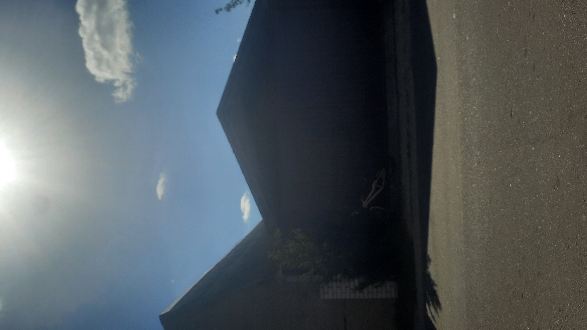 The image depicts a street scene in Shklov, Belarus, under a bright, hazy sun. In the foreground, a paved road occupies the lower portion of the frame. The middle ground features the side of a dark, angular building with a gabled roof. To the left, a smaller, lighter-colored section of the same building is visible, with a brick wall element. A tree or large bush with green foliage grows near the corner of the buildings, partially obscuring a parked bicycle that is lying on its side in the shadows. In the background, the sky is a clear blue with a few wispy white clouds and the sun is prominently visible at the top, creating a glare that washes out some of the sky's details. The overall impression is an outdoor, possibly industrial or storage area, on a sunny day.