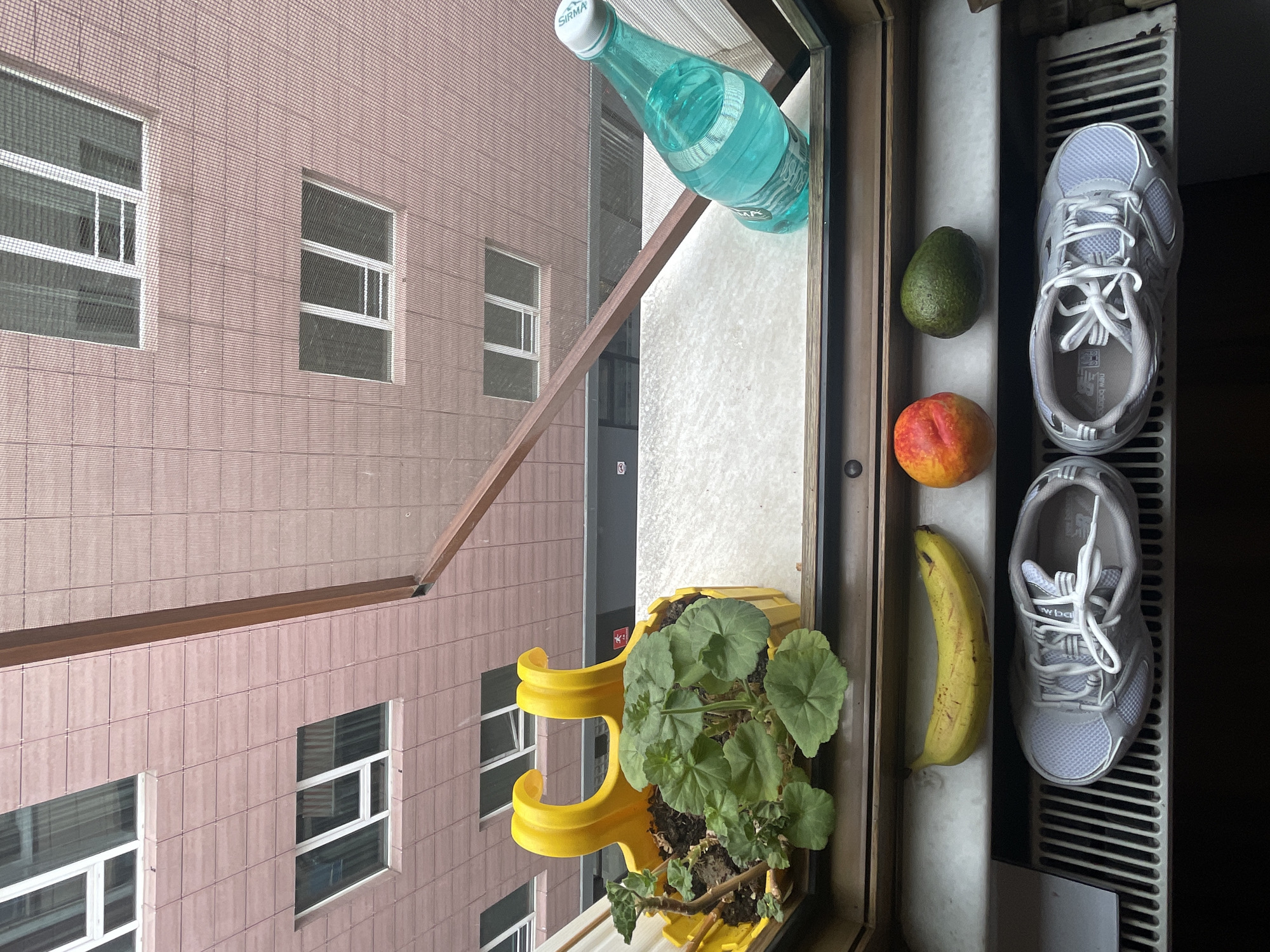 The image depicts a windowsill in Kadıköy, Turkey, overlooking a building with a pink tiled facade and multiple windows. On the windowsill, from left to right, are a yellow banana, a peach, and an avocado. Below the windowsill, on a radiator, are a pair of grey New Balance sneakers. To the right, a potted plant with green leaves sits in a bright yellow planter. Further to the right on the windowsill is a turquoise bottle of "Sirma" brand sparkling water. The scene appears to be an outdoor setting, possibly a balcony or an open window.