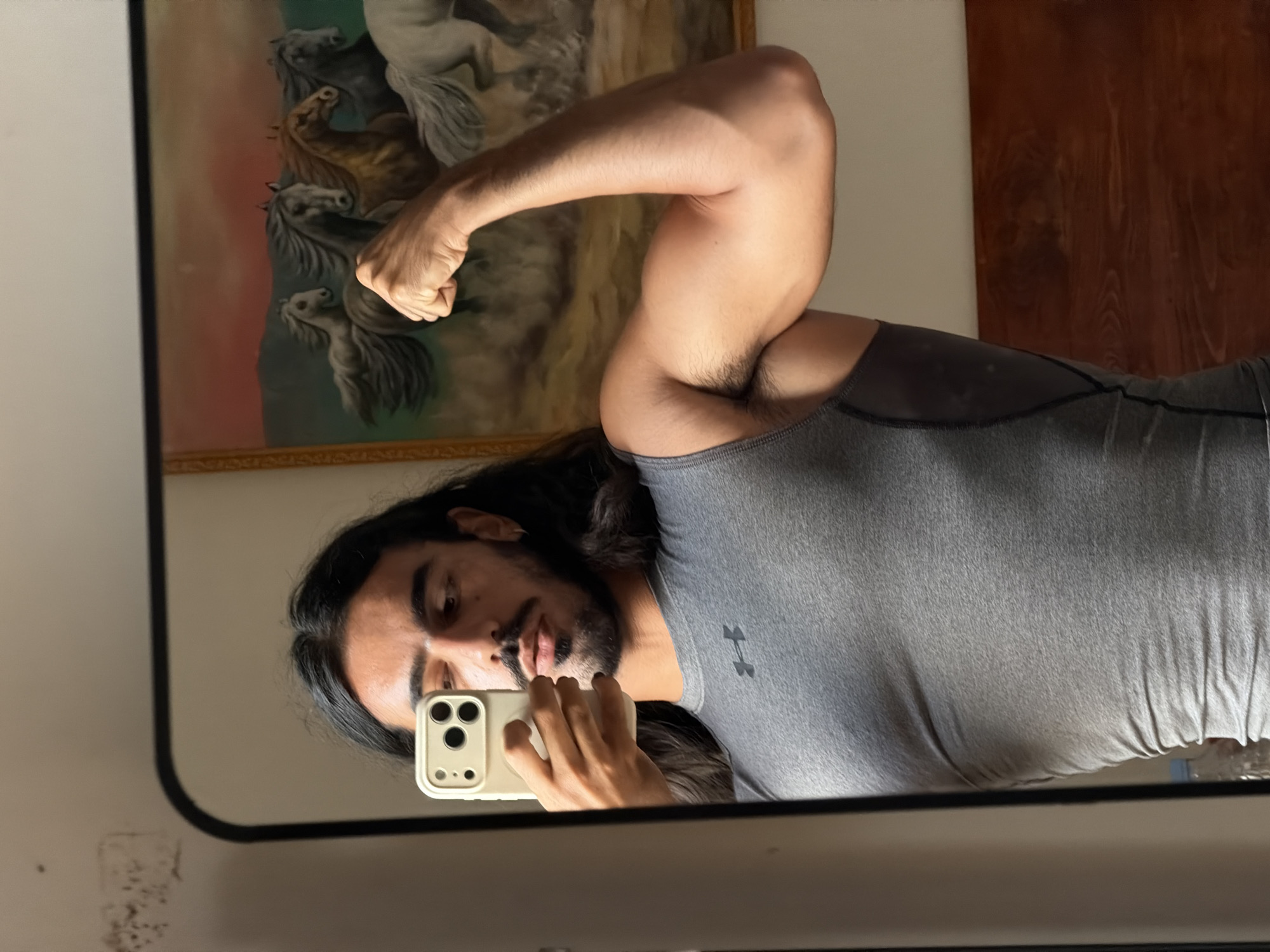 In a dimly lit room, a man with long dark hair and a beard is taking a mirror selfie. He is wearing a grey Under Armour compression shirt and flexing his right bicep. The mirror reflects a painting of horses running in a field against a sunset sky. The room appears to be a bedroom, with a wooden headboard visible in the background.