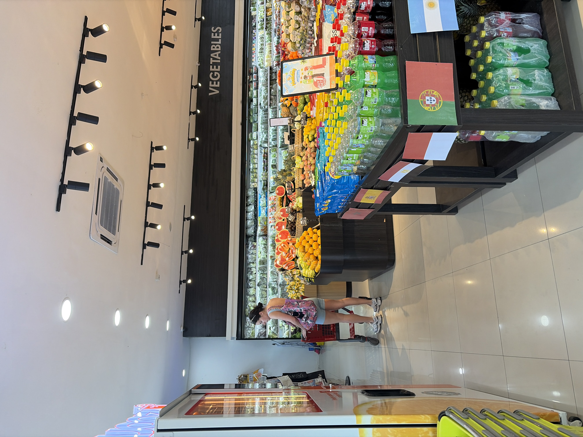 This image depicts the interior of a brightly lit supermarket in North Kuta, Indonesia. In the foreground, a woman in a pink patterned tank top and light blue shorts stands beside a red shopping cart, facing away from the camera. She is wearing white athletic shoes and has her hair pulled back. To her left, a vending machine with orange and yellow graphics is partially visible.
The background reveals a well-stocked produce section with rows of fresh fruits and vegetables displayed on shelves and in baskets. A large sign above the produce reads "VEGETABLES". In front of the produce section, a display table holds numerous bottles of beverages, some in green packaging and others in red. Several small national flags, including those of Spain, Egypt, Portugal, and Argentina, are attached to the display table. The floor is tiled with large, light-colored square tiles. The ceiling features recessed lighting and track lighting fixtures. The overall environment suggests a clean and organized retail space.