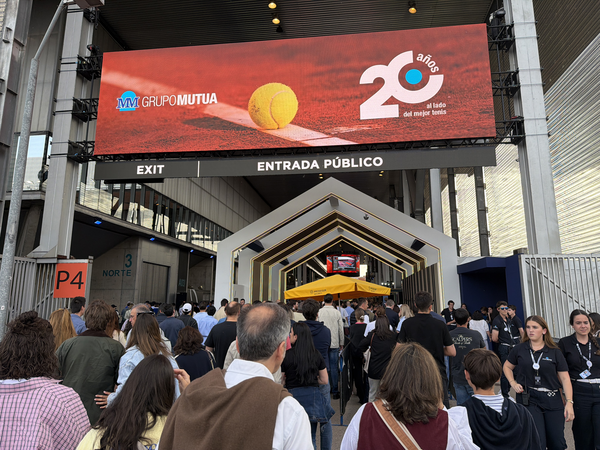 The image shows a crowd of people entering the Mutua Madrid Open tennis tournament. Above the entrance is a large digital display showing an advertisement for Grupo Mutua, celebrating 20 years of being involved with "the best tennis". The entrance is marked "ENTRADA PÚBLICO" and there is also an "EXIT" sign. A sign for "P4" and "3 NORTE" is visible near a gate. Security personnel wearing black shirts with lanyards are visible on the right. A yellow umbrella with "PROSEGUR SECURITY" printed on it is positioned in front of the crowd. The scene is outside a stadium or venue in Madrid, Spain.