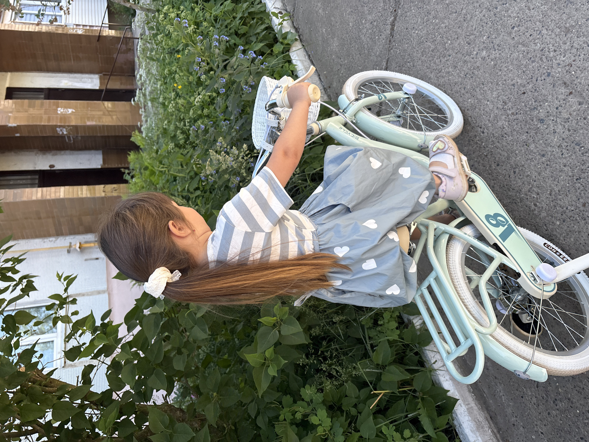 A young girl with long brown hair tied back with a white scrunchie is sitting on a mint green bicycle with white tires. She is wearing a striped t-shirt and a light blue skirt with white hearts. The bicycle has a white basket on the front. The girl is on a paved path next to a building with brown and white vertical accents. Green foliage and plants are visible in the background. The overall scene suggests an outdoor recreational activity.