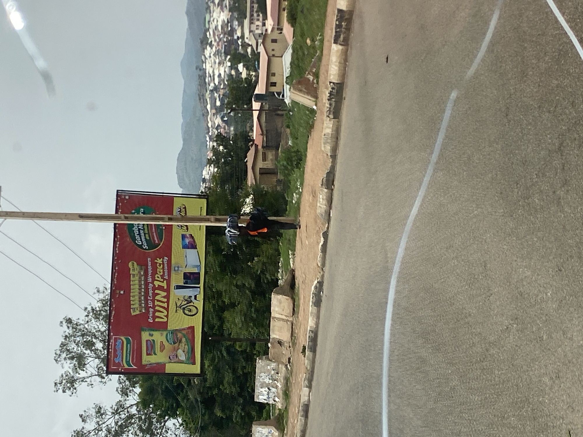 A billboard for Indomie noodles is visible on the left side of the frame. The billboard has a red background with yellow lettering. It features an image of an Indomie noodle packet and text that reads "Bring 10 Empty Wrappers WIN 1 Pack Instantly". Below the text, there are illustrations of a bicycle, a gaming console, and a refrigerator. A person is walking on the sidewalk on the right side of the billboard, carrying a bundle of items on their head. The person is wearing a black shirt and orange vest. The scene is set in Jos, Nigeria, with a hilly landscape and buildings visible in the background. The foreground shows a road with white markings. Concrete barriers are also present along the sidewalk. The sky is overcast.