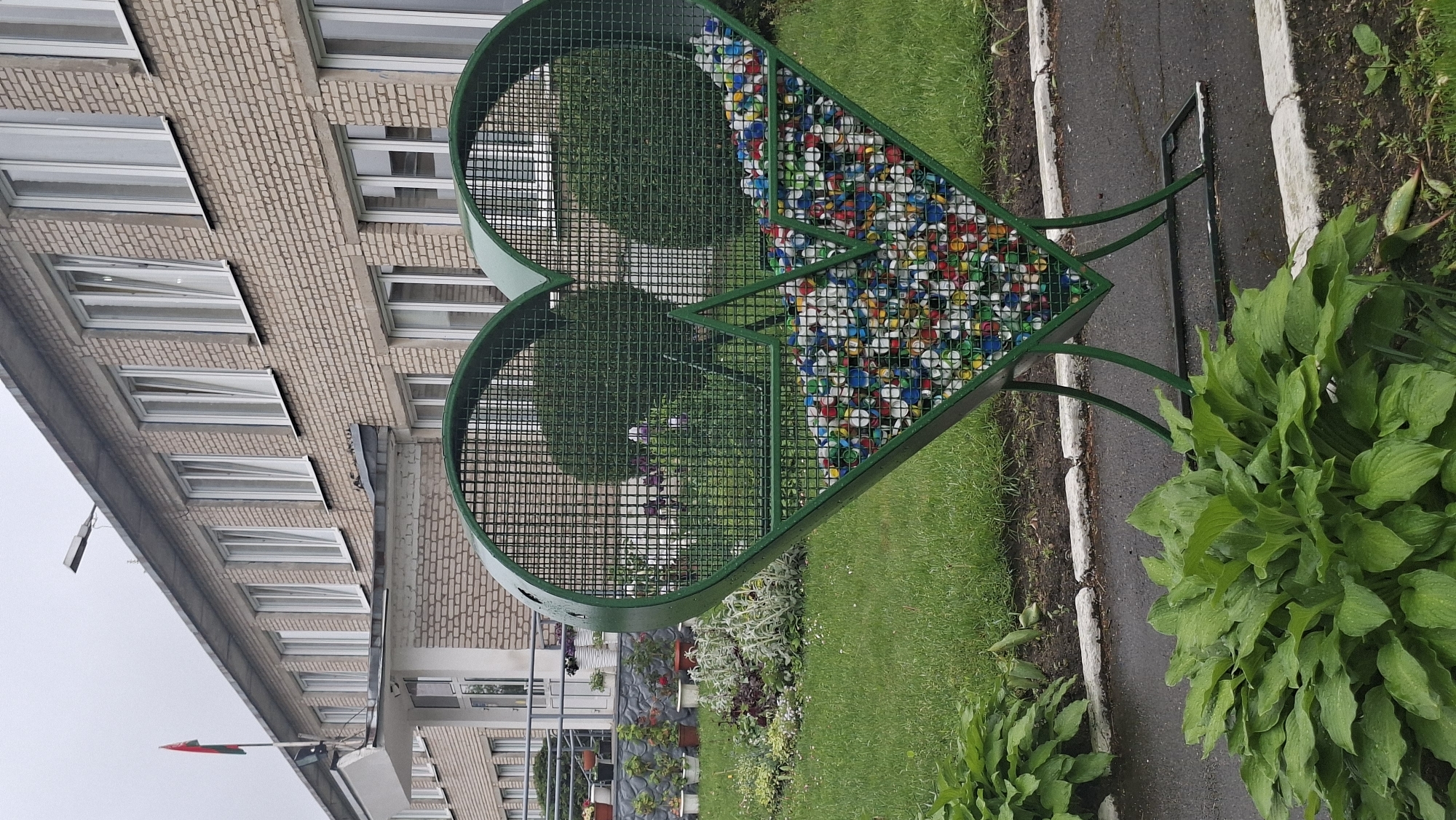 The image shows a green heart-shaped wire sculpture filled with colorful plastic bottle caps. The sculpture is located on a grassy area next to a sidewalk in Забалоцкі сельскі Савет, Belarus. A brick building with numerous windows is visible in the background. Some plants and flowers are present around the sculpture.