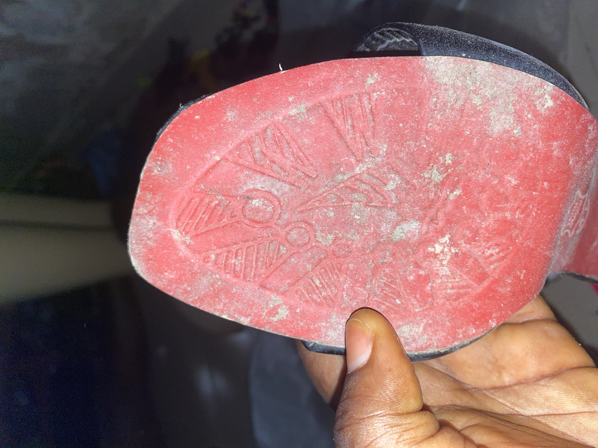 The image shows a close-up view of the red sole of a shoe, held by a person's hand. The sole has a patterned design and is covered in a light layer of dust. The background is dark and out of focus, with hints of what appear to be indoor surroundings. The scene is static and does not depict any specific activity. No discernible location within Jauro Yinu, Nigeria can be identified from the provided image.