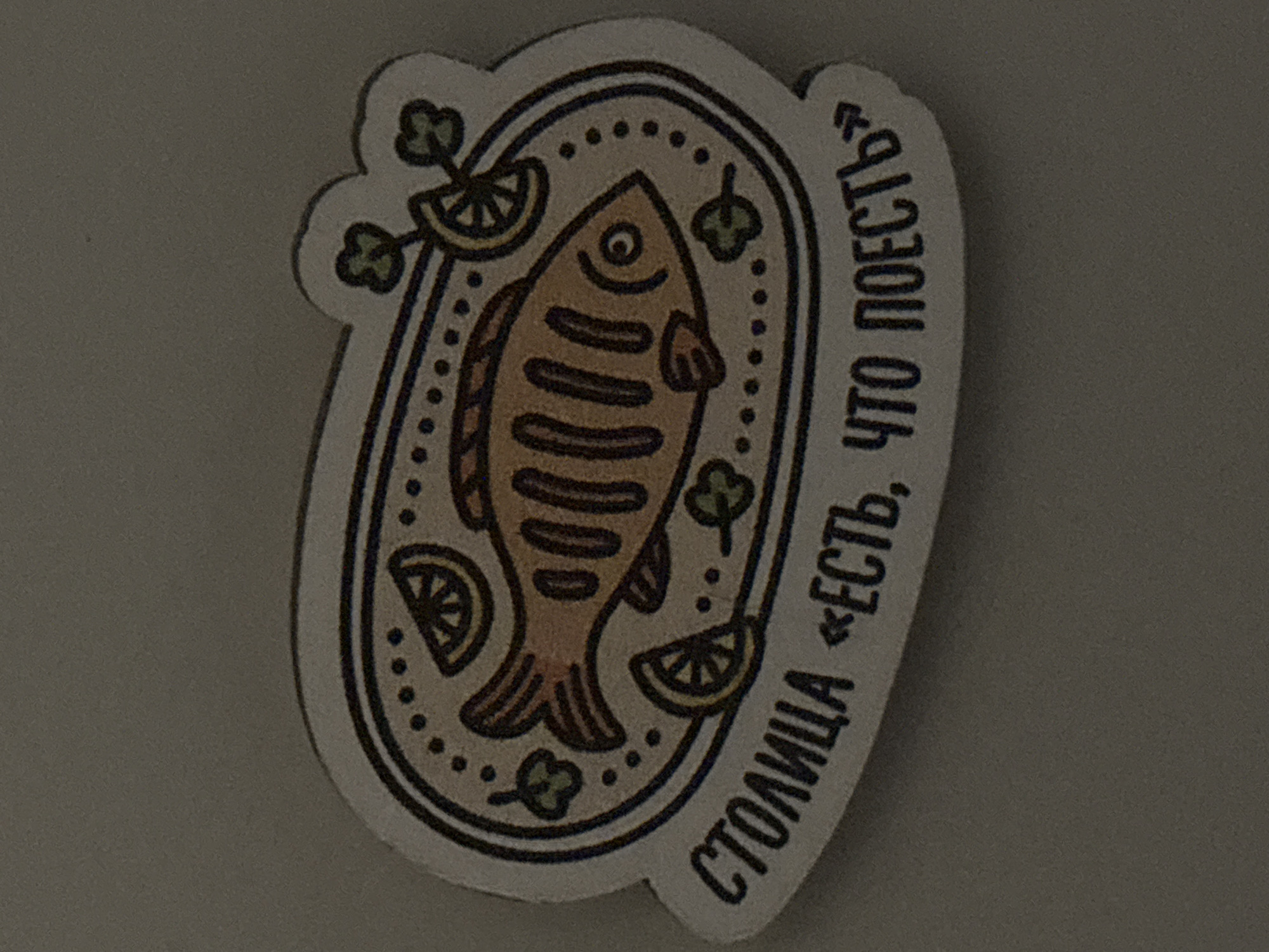 The image displays a close-up of a sticker featuring a stylized illustration of a fish on an oval plate. The fish, colored in shades of brown, is depicted with simple lines indicating scales and fins, and a distinct circular eye. It is garnished with several yellow lemon wedges and small green sprigs, possibly parsley or dill, arranged around it on the plate. The plate itself is outlined with a dotted pattern inside a thicker, dark border.
Along the bottom and right edges of the sticker, there is Cyrillic text. The text reads: "СТОЛИЦА «ЕСТЬ, ЧТО ПОЕСТЬ»". This translates to "CAPITAL «THERE IS, WHAT TO EAT»" or "CAPITAL «SOMETHING TO EAT»," suggesting a slogan or a name, possibly for a restaurant or food-related establishment. The sticker has a light background, and it is placed against a plain, dark gray surface. The style of the illustration is reminiscent of a simple, hand-drawn or folk art aesthetic. No specific time of day, weather, or emotions are conveyed by the image. While the location context is Krasnoyarsk, Russia, there are no visual cues within the image to confirm this.