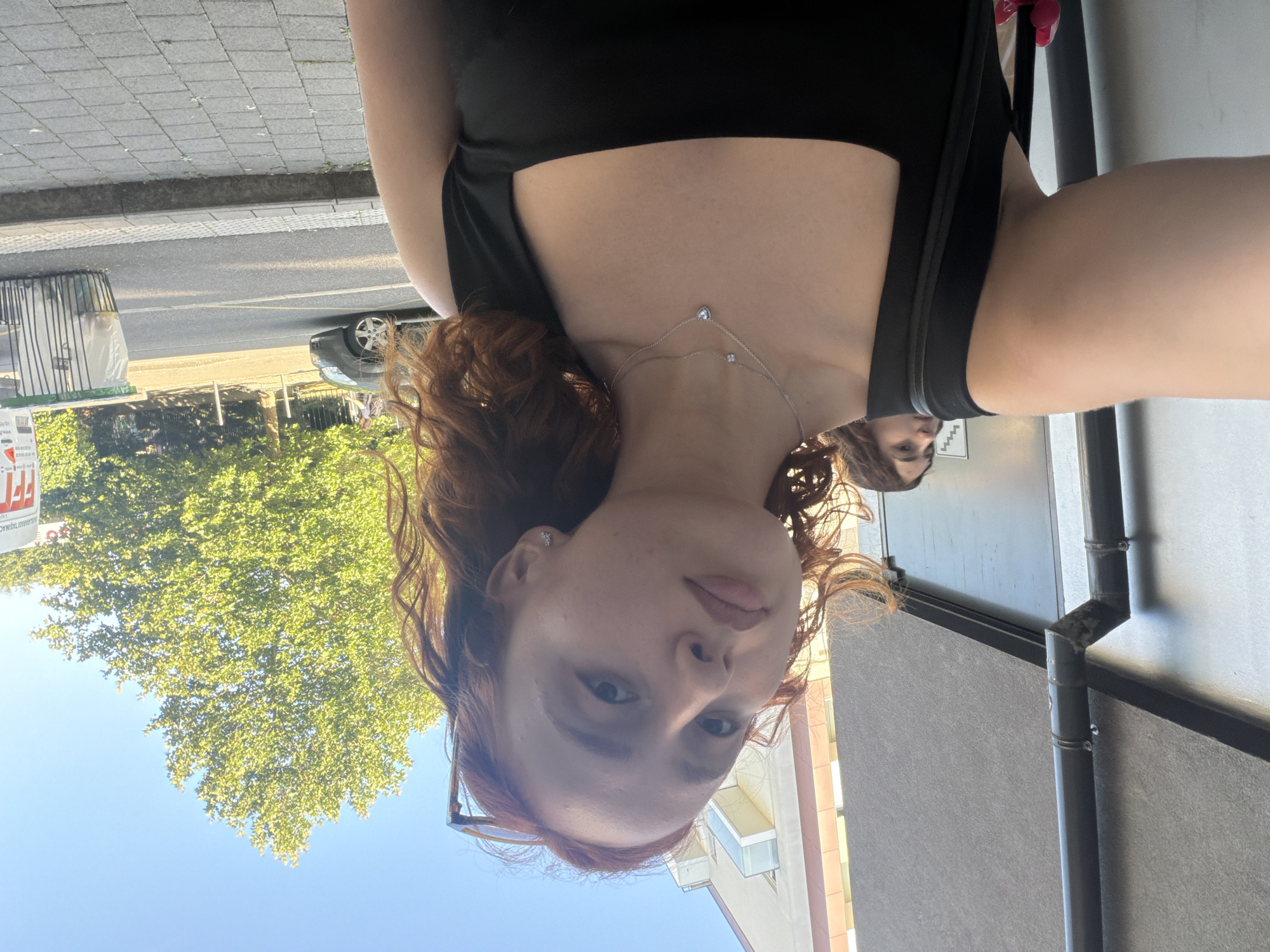 A young woman with shoulder-length, wavy auburn hair is taking a selfie outdoors. She is wearing a black tank top and two delicate silver necklaces. Her gaze is directed towards the camera. In the background, slightly out of focus, another young woman with brown hair is visible. To the left, a gray drainpipe runs down the side of a building with a textured gray facade. The background also shows a street with a car, a large green tree, and a trash can. The setting appears to be a street scene in the city of Strasbourg, France, on a bright, sunny day.