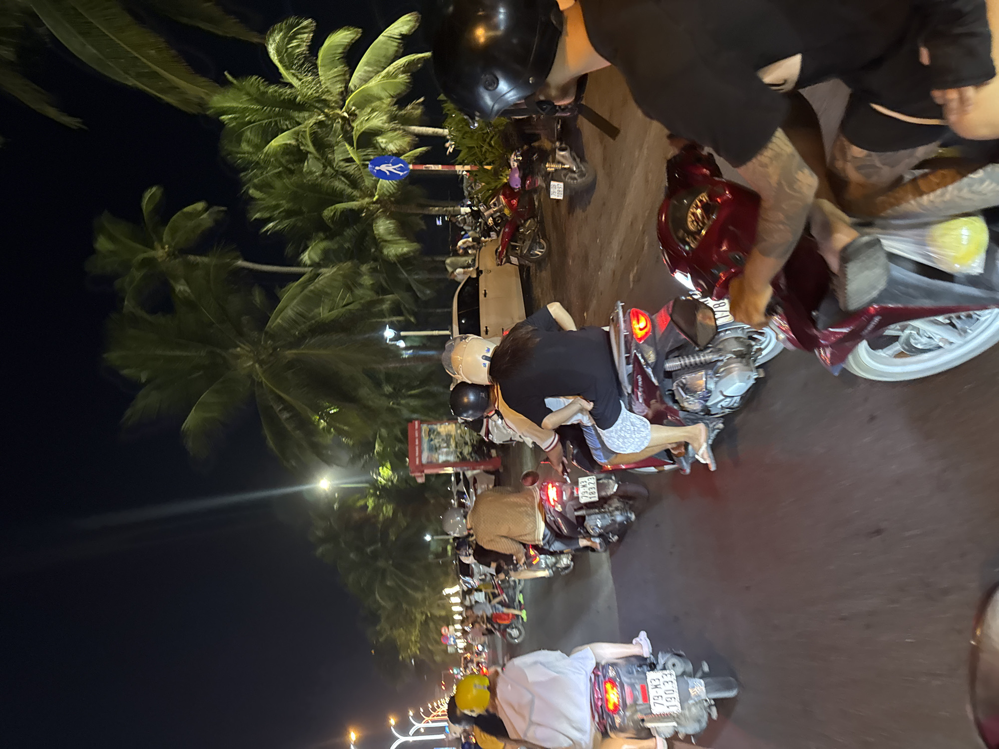 This image captures a bustling nighttime street scene in Nha Trang Ward, Khánh Hòa Province, Vietnam, confirmed by the '79' prefix on visible motorbike license plates. The environment is illuminated by artificial streetlights and vehicle lights, revealing a clear, dry night.
The foreground and midground are dominated by numerous motorbikes and scooters, primarily occupied by one or two riders each, mostly wearing helmets. From left to right:
* A scooter with license plate **79-M3 190.33** carries two individuals; the rider wears a yellow helmet and a light-colored long-sleeved top.
* Another scooter, with license plate **79-M3 183.23**, has a single rider in a light-colored helmet and a brown patterned jacket.
* On the far right, a maroon scooter, partially visible, has a rider with heavily tattooed arms and a black t-shirt, wearing a black helmet, and holding a clear plastic bag containing a yellow liquid.
Numerous other motorbikes and a white car are visible further down the street. The road surface is asphalt. Abundant tall palm trees with green fronds line both sides of the street, characteristic of a coastal setting. A blue circular street sign displaying a white pedestrian icon is visible on the right, affixed to a pole. A red rectangular kiosk-like structure is also present on the right side of the road further back.