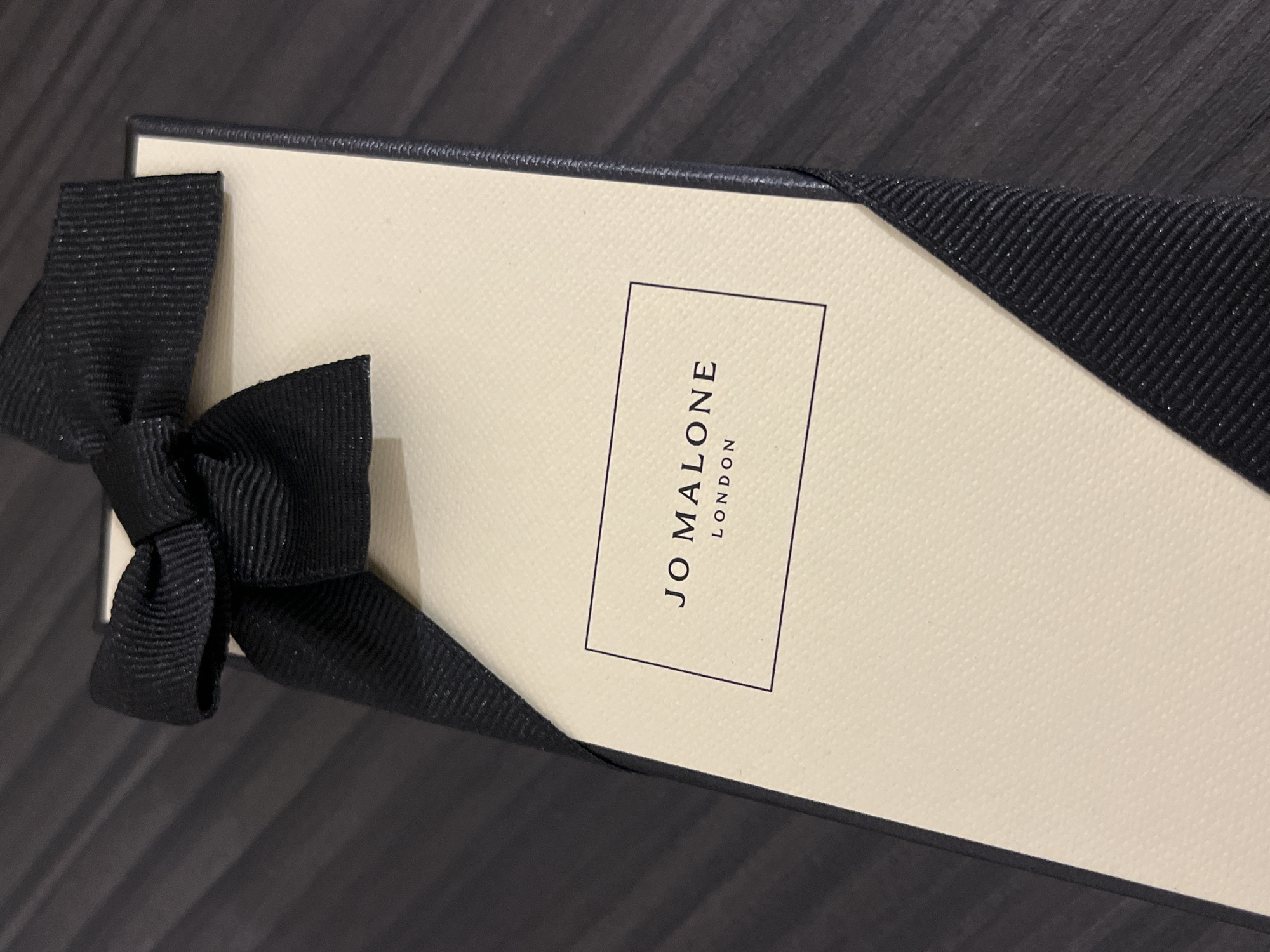 A close-up shot shows a Jo Malone London box on a dark wood-grained surface. The box is light beige with a black border and a black ribbon tied into a bow on the left side. The "JO MALONE LONDON" logo is centered in a black rectangle on the box's surface. The box is at a slight angle. No people are visible in the image. The location is unknown.
