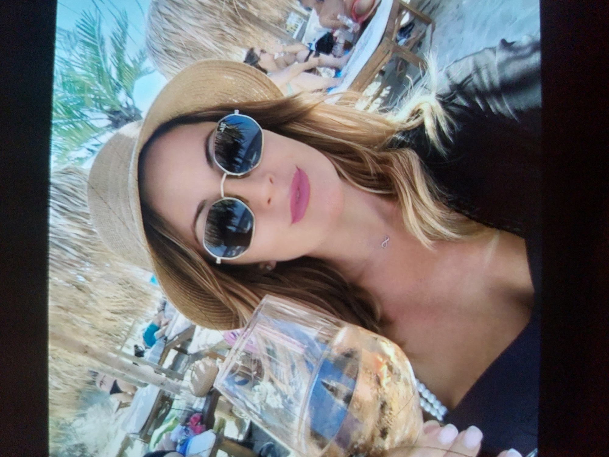 A woman is captured in a selfie at a beach in Tsarevo, Bulgaria. She is in the foreground, looking directly at the camera, holding a clear glass filled with an amber-colored drink, possibly wine or a cocktail. She has long, blonde hair, wears a light straw hat, and round, dark sunglasses. Her lips are painted with pink lipstick, and an infinity symbol necklace is visible on her chest. She is wearing a dark top, possibly a swimsuit or dress.
The background reveals a sunny beach scene with a clear blue sky and several palm trees. Other individuals are visible in the background, out of focus, relaxing on sun loungers or beach beds under thatched umbrellas or canopies, typical of a beach resort or bar setting. The overall atmosphere suggests a relaxed, leisurely day at the coast. The image is bright, indicating it's daytime.