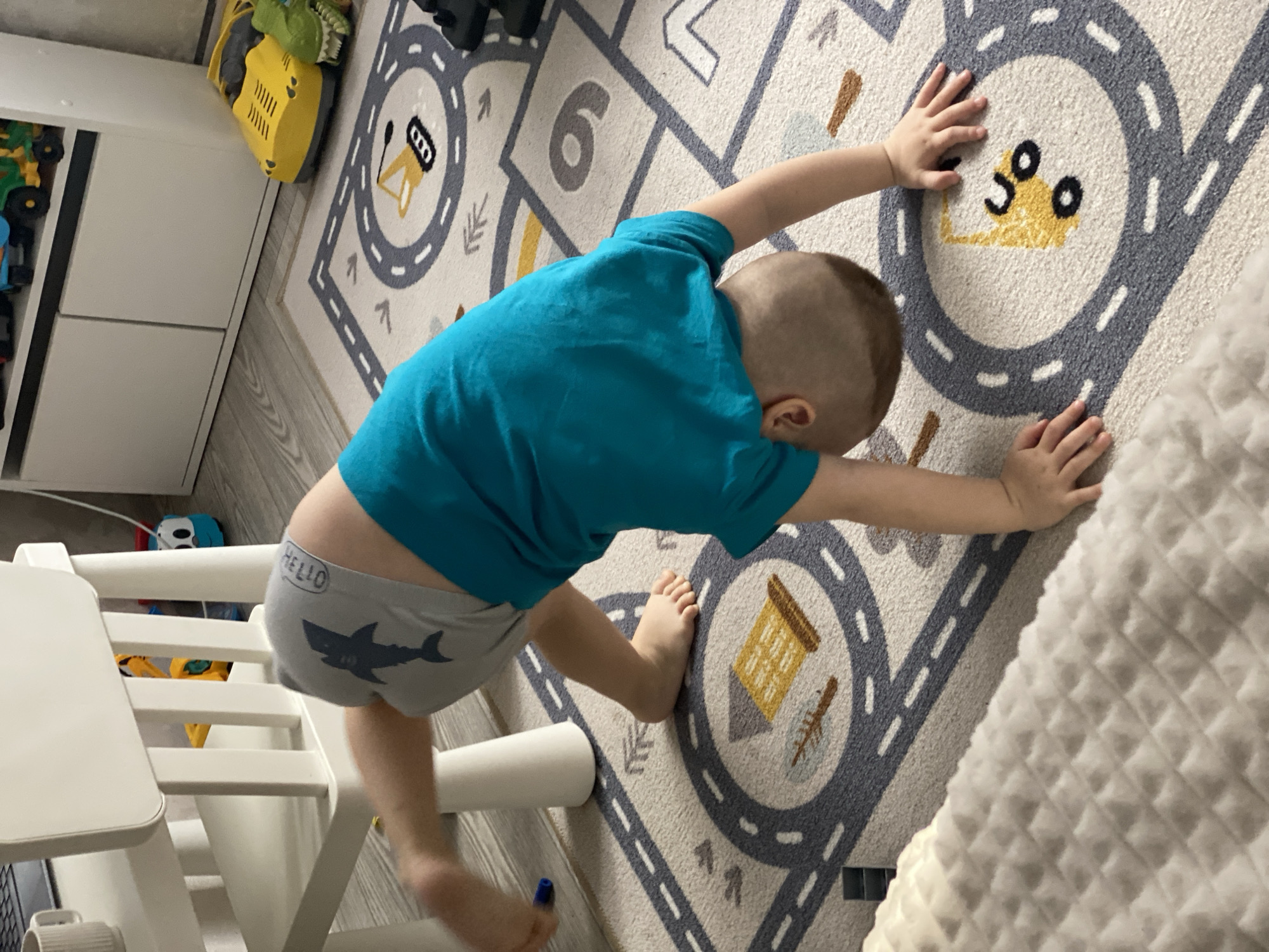 A child is positioned with their body on a rug, reaching out with their hands. The child is wearing a blue shirt and gray underwear with a shark design on the rear. The rug features a pattern resembling a road with various numbered circles. A white chair is situated nearby, and in the background, there is a white cabinet with yellow and green toys. The location cannot be determined as Sochi, Russia, based on the image.