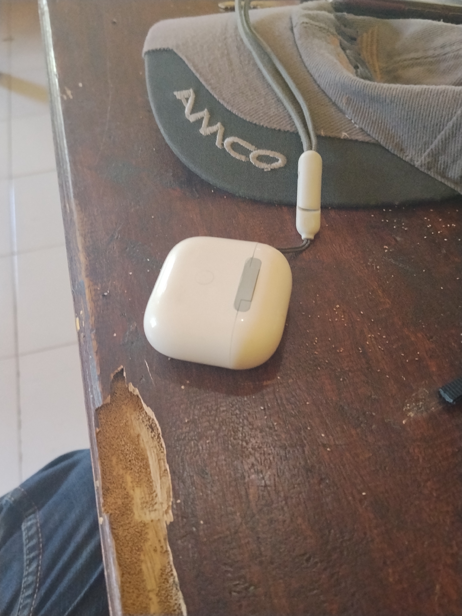 The image shows a close-up, high-angle view of a weathered wooden table. On the table rests a white wireless earbud case with a gray button on its surface. The earbud case is connected by a gray string to a white cylindrical object. Next to the earbud case is a dark green cap with a gray brim, embroidered with the white lettering "AMCO". The cap is partially covering a portion of the wooden table, which exhibits significant wear and tear, with peeling veneer exposing the lighter wood underneath. In the bottom left corner of the image, a portion of dark blue denim fabric is visible, suggesting the presence of a person wearing jeans. The surface of the table is scattered with small, light-colored particles. The background is blurred, but appears to be an indoor setting with a tiled floor. The scene is set in Laok Jangjang, Indonesia.