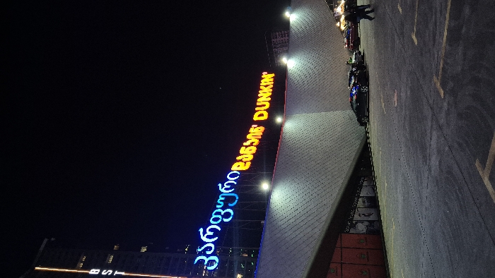 A vertically oriented night shot showcases a Dunkin' Donuts sign, illuminated in orange, above a Georgian script sign in blue and orange. The building's facade is constructed with a patterned gray material. Adjacent to the building is a parking lot with parked cars and pedestrian figures visible. The darkness of the sky contrasts with the lit signs and parking area.