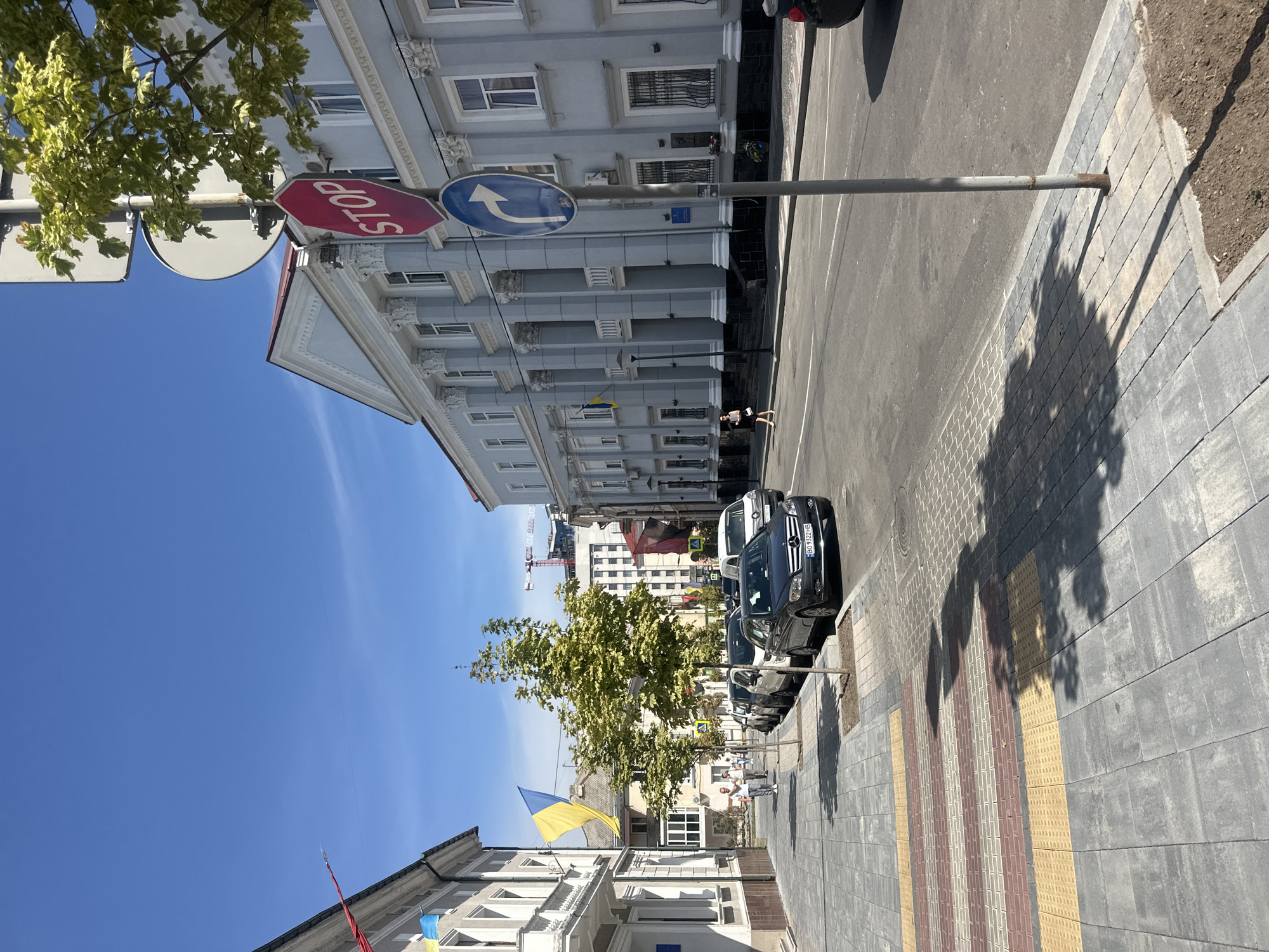 This is a street scene in Ternopil, Ukraine, on a sunny day with a clear blue sky. A paved sidewalk is in the foreground, featuring a pedestrian crossing with yellow and red tactile paving. Several cars are parked along the street, including a dark sedan with license plate B09322HB in the foreground. A woman in a dark dress and light-colored shoes is walking on the sidewalk. To the right of the street, a large, light blue building with columns and arched windows stands prominently. A red octagonal "STOP" sign and a blue circular sign with a right-pointing arrow are mounted on a pole in front of the building. Ukrainian flags are visible on the buildings and on poles along the street. Trees with green foliage line the sidewalk. In the background, construction cranes and other buildings can be seen. The overall impression is one of an urban environment with some trees and pedestrian activity.