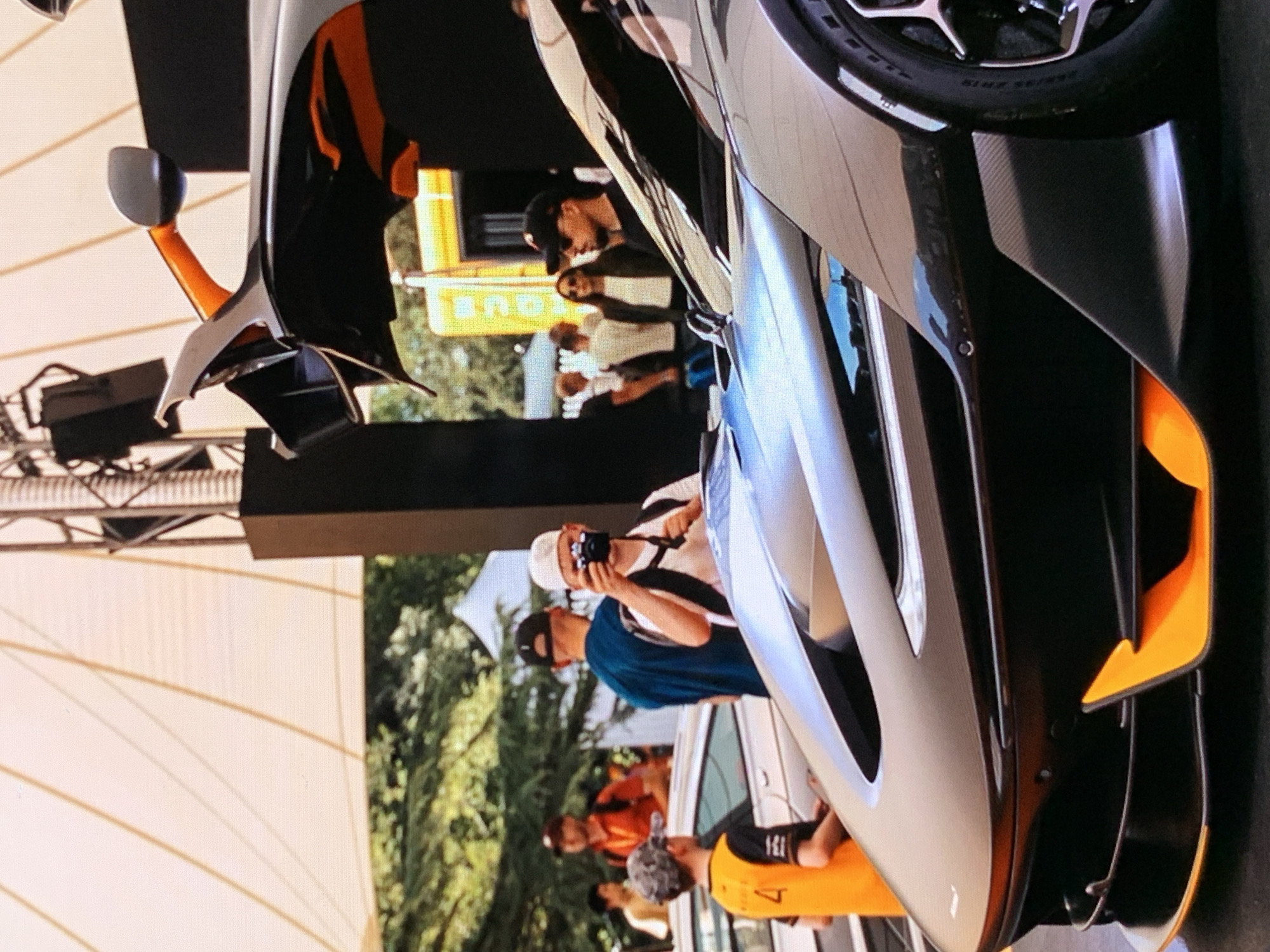 A dark McLaren sports car with orange accents is prominently displayed at what appears to be an outdoor event, possibly a car show. The car's dihedral doors are open, revealing a portion of its interior. In the foreground, the front of the car is visible, featuring aggressive styling with orange aerodynamic elements. Behind the car, a group of people are gathered. One individual in the mid-ground is holding a camera and taking a picture. Another person, wearing an orange vest with the number '4', is standing to the left. Further in the background, trees and a tent-like structure suggest an outdoor venue. The lighting indicates it is daytime, possibly under overcast skies or a large canopy. The overall setting is an organized public gathering centered around the display of the luxury vehicle.