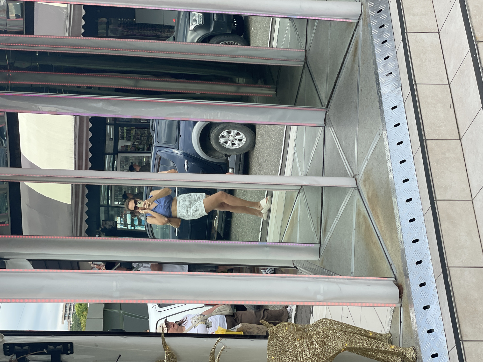 The image shows a woman taking a selfie in a funhouse mirror attraction, likely in Sochi, Russia. The woman is wearing a blue crop top and white floral shorts, and she is holding a smartphone to take the picture. In the background, a dark blue SUV is parked, and some buildings are visible. The funhouse mirrors are arranged vertically, creating multiple reflections of the woman and the surrounding environment. The ground is tiled and there is a metal ramp with drainage holes in the foreground. A gold sequined reindeer decoration is partially visible on the left side of the image.