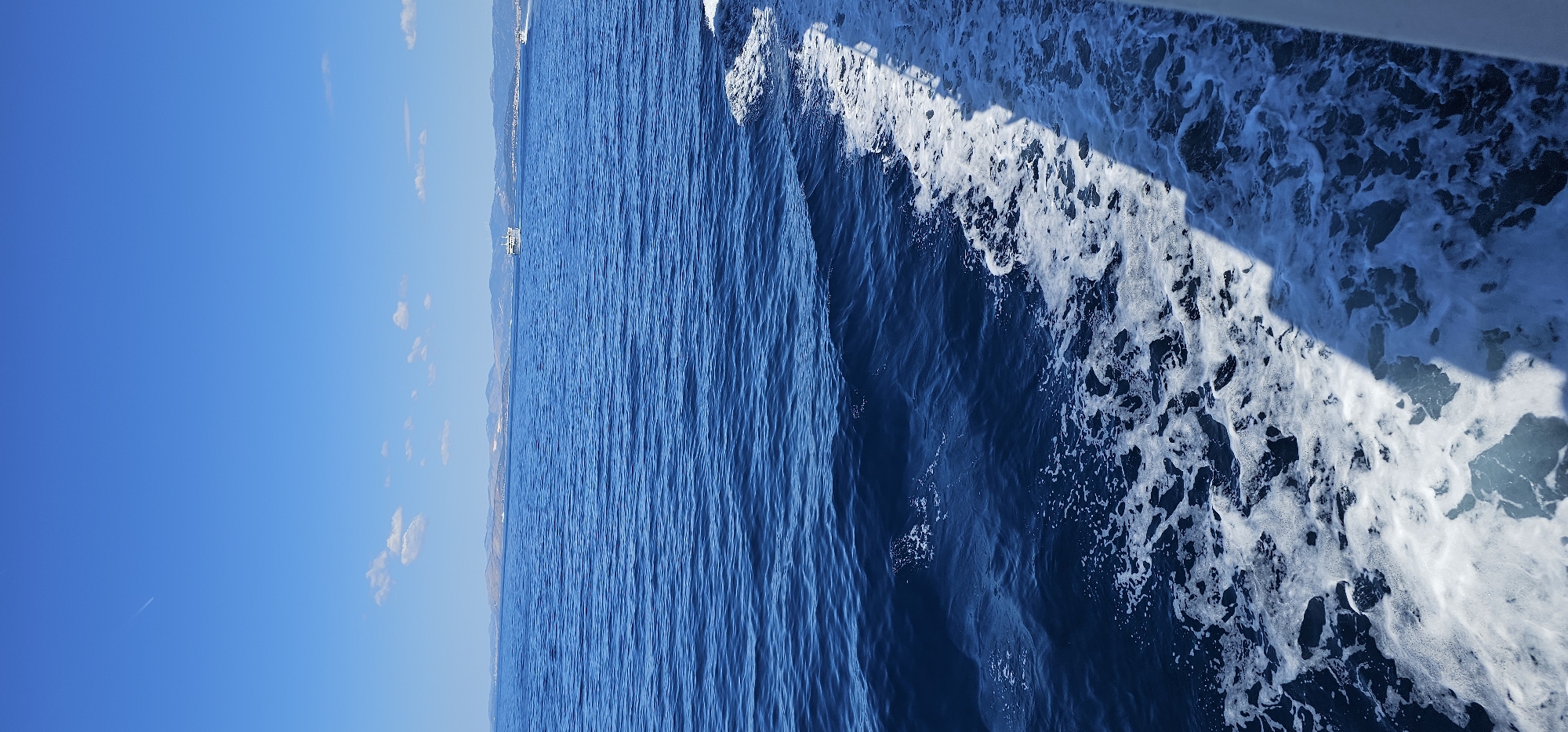 This is a medium shot, horizontal-format image taken from a boat moving through the sea. The water is a deep, rich blue, with visible waves and white foam generated by the boat's passage. A shadow from the boat's structure falls across the water on the right side of the frame, highlighting the texture of the churning sea. In the distance, a coastline is visible, lined with mountains. A white ferry or larger boat is also visible on the horizon. The sky is a clear, bright blue with a few small, scattered clouds. A faint contrail from an airplane is visible in the upper left portion of the sky. The overall impression is of a bright, sunny day at sea, with the camera looking towards the distant land. The scene evokes a sense of travel and a peaceful day outdoors. There is no visible text.