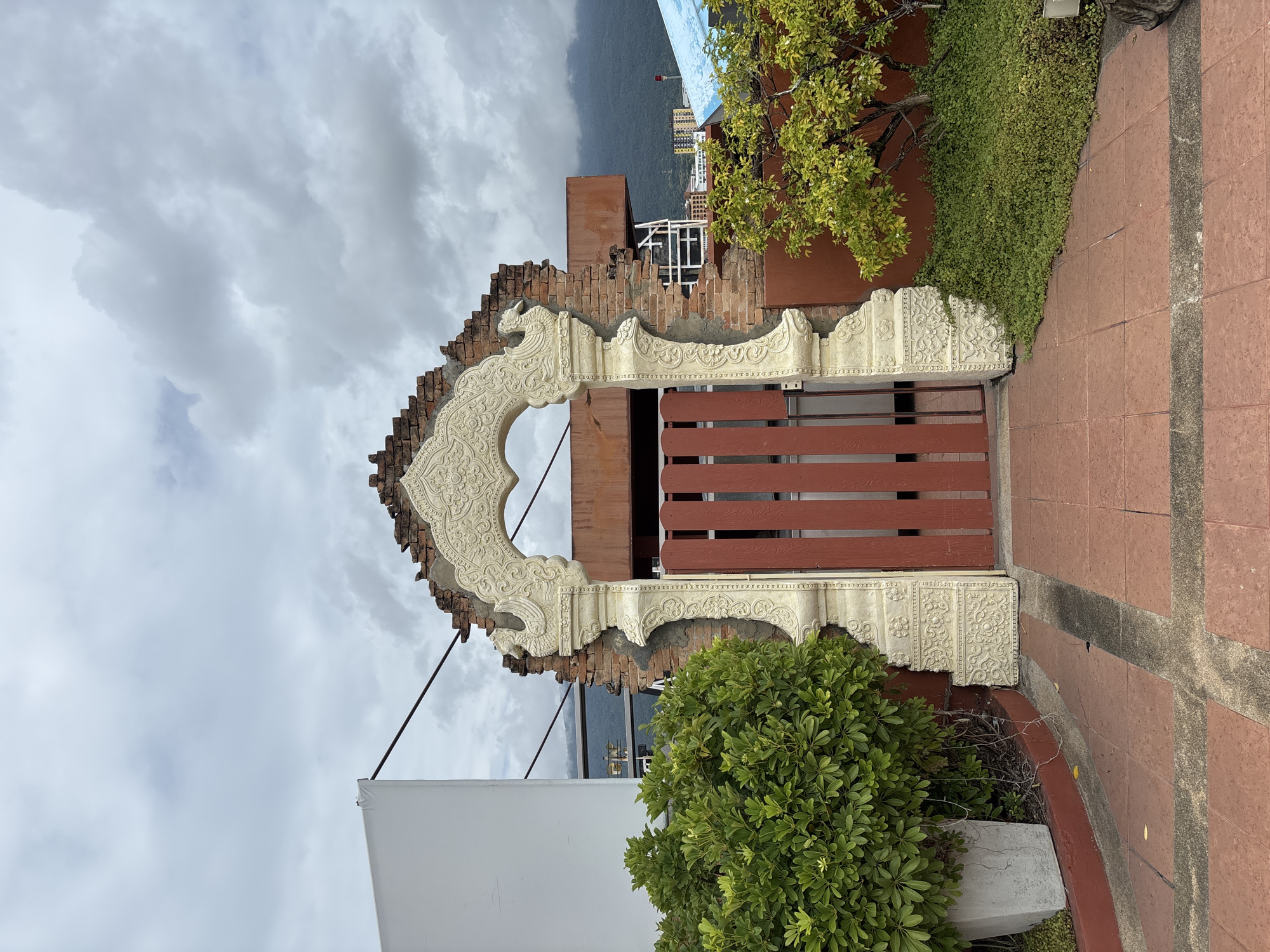 In Chiang Mai City Municipality, Thailand, an ornate, ruined archway stands as a focal point, framing a red wooden gate. The archway is crafted from a pale, decorative material, with intricate carvings of mythical creatures and floral patterns adorning its surface. Behind the archway, exposed brickwork reveals the weathered remnants of a structure, hinting at past grandeur. To the left of the archway, a large, lush green bush is contained within a concrete planter, its leaves dense and vibrant. The ground leading up to the archway is paved with reddish-brown tiles, laid in a geometric pattern. To the right of the archway, a small tree with light green foliage adds a touch of natural beauty to the scene, with a carpet of low-lying green ground cover spreading beneath it. The background reveals a cityscape with buildings of various heights and architectural styles, nestled against a backdrop of misty, green mountains under a cloudy sky. The overall atmosphere is one of contrast, where the delicate artistry of the archway juxtaposes with the decaying brickwork and the urban and natural landscape beyond.
