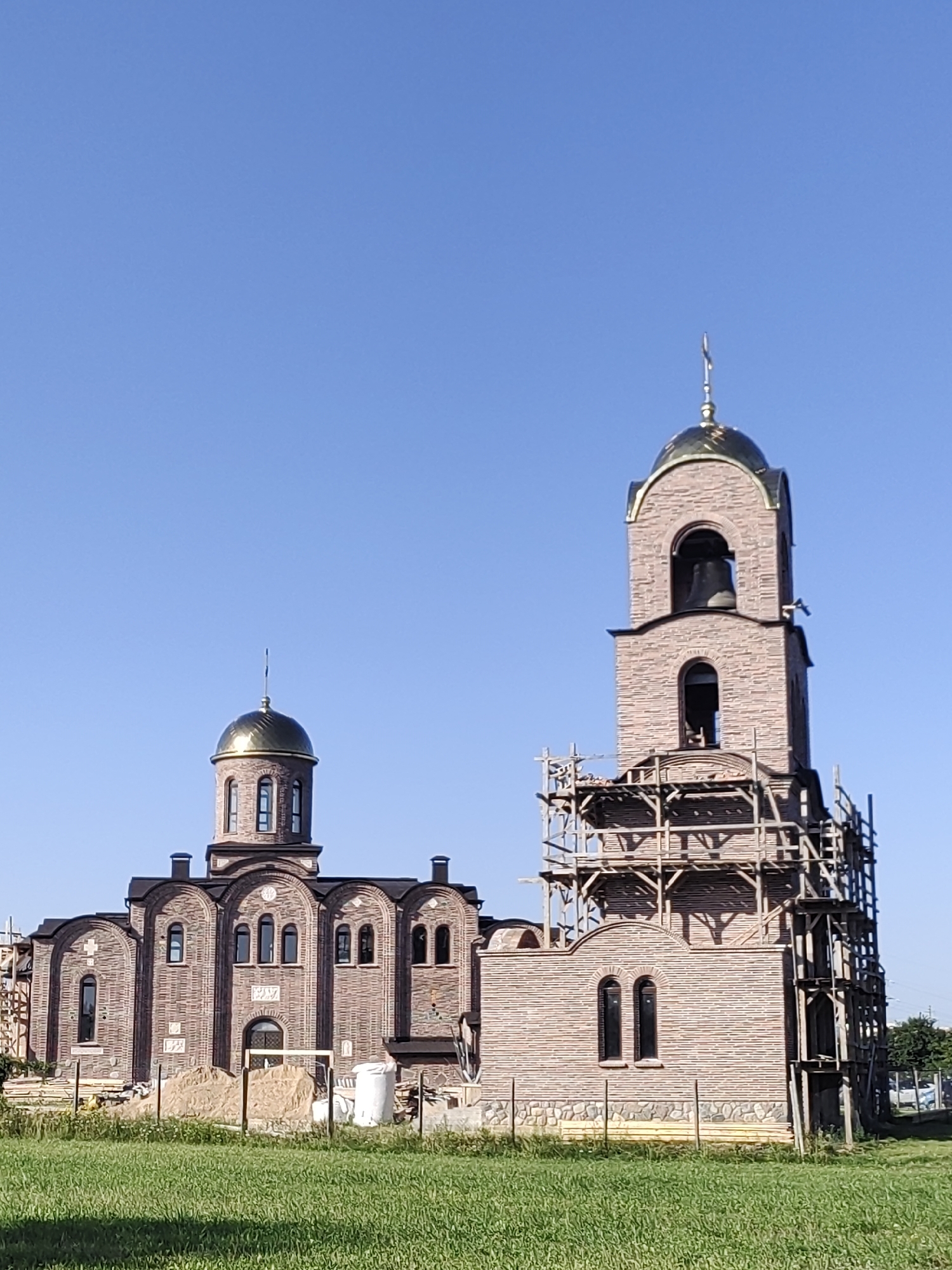 A brick church building is under construction with scaffolding around the bell tower. The church has a golden dome and a bell in the tower. The building is surrounded by a grassy field and a clear blue sky.