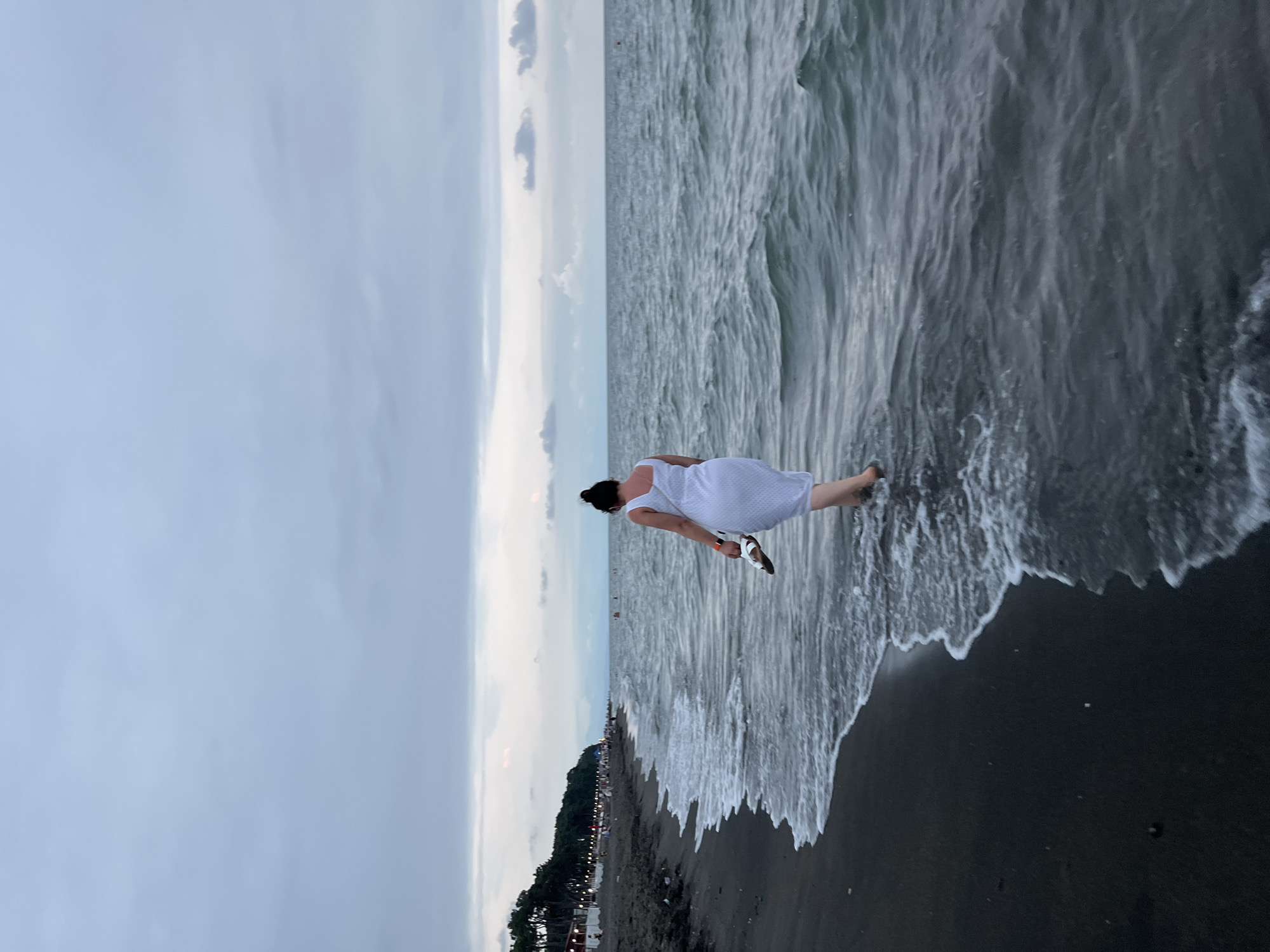 A woman in a white dress is walking into the sea at Ureki, Georgia. She is holding a pair of white sandals in her right hand. The water is dark and choppy, with white foam washing onto the black sand beach. In the background, there are trees and a few other people on the beach. The sky is overcast and a pale gray. The overall mood of the image is serene and peaceful.