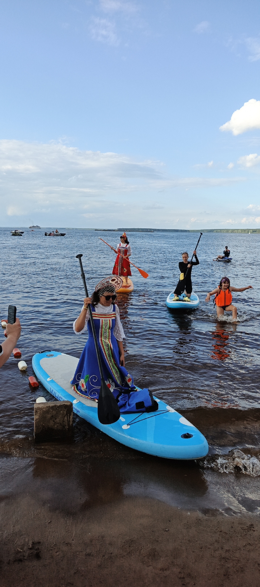 On a sunny day, a group of people are paddleboarding on the Kama River in Chaikovsky, Russia. The foreground is dominated by a large, blue paddleboard with a woman dressed in a traditional blue and white folk costume. She is holding a paddle and looking towards the camera. To her left, another person's hand holding a phone is visible, suggesting they are taking a picture. In the background, several other paddleboarders can be seen, some in colorful outfits. Further in the distance, boats and a large ship are on the river, and a tree-lined shore is visible. The sky is a clear blue with scattered white clouds. The water is calm and reflects the blue sky. The scene appears to be part of a cultural event or celebration.