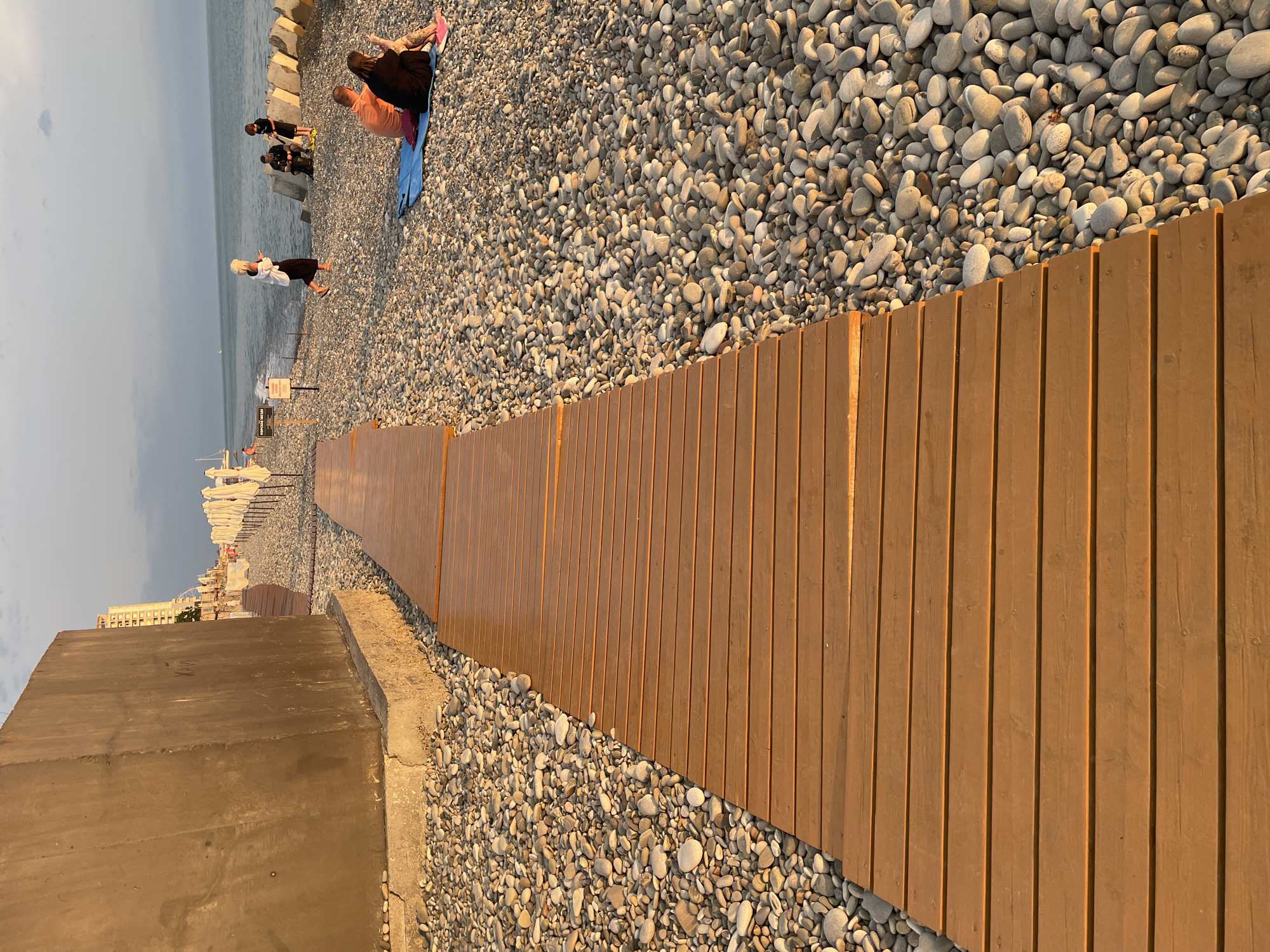 A wooden walkway traverses a pebble beach in an outdoor setting with the sea visible in the background. The walkway is made of wide, plank-like boards with a warm, orange-brown hue. It leads from the foreground and curves to the right, disappearing out of frame. The beach is covered in smooth, grey and white pebbles of varying sizes.
Several people are present on the beach. In the middle ground, a woman in a white top and black skirt is walking, holding a phone. Nearby, two figures, a man and a boy, are seated on the pebbles. Further in the background, a couple is sitting on a blue mat, and another person is seated beside them. Some white beach umbrellas are lined up along the shore.
To the left of the walkway, a large, rough concrete structure stands prominently, its base resting on the pebble beach. In the far background, a tall building is visible, suggesting an urban or developed coastal area. The sky above is partly cloudy, and the setting sun casts a warm glow over the scene, creating long shadows and highlighting the textures of the pebbles and the wood. The overall impression is a serene beach environment with people enjoying a late afternoon or early evening by the sea.
