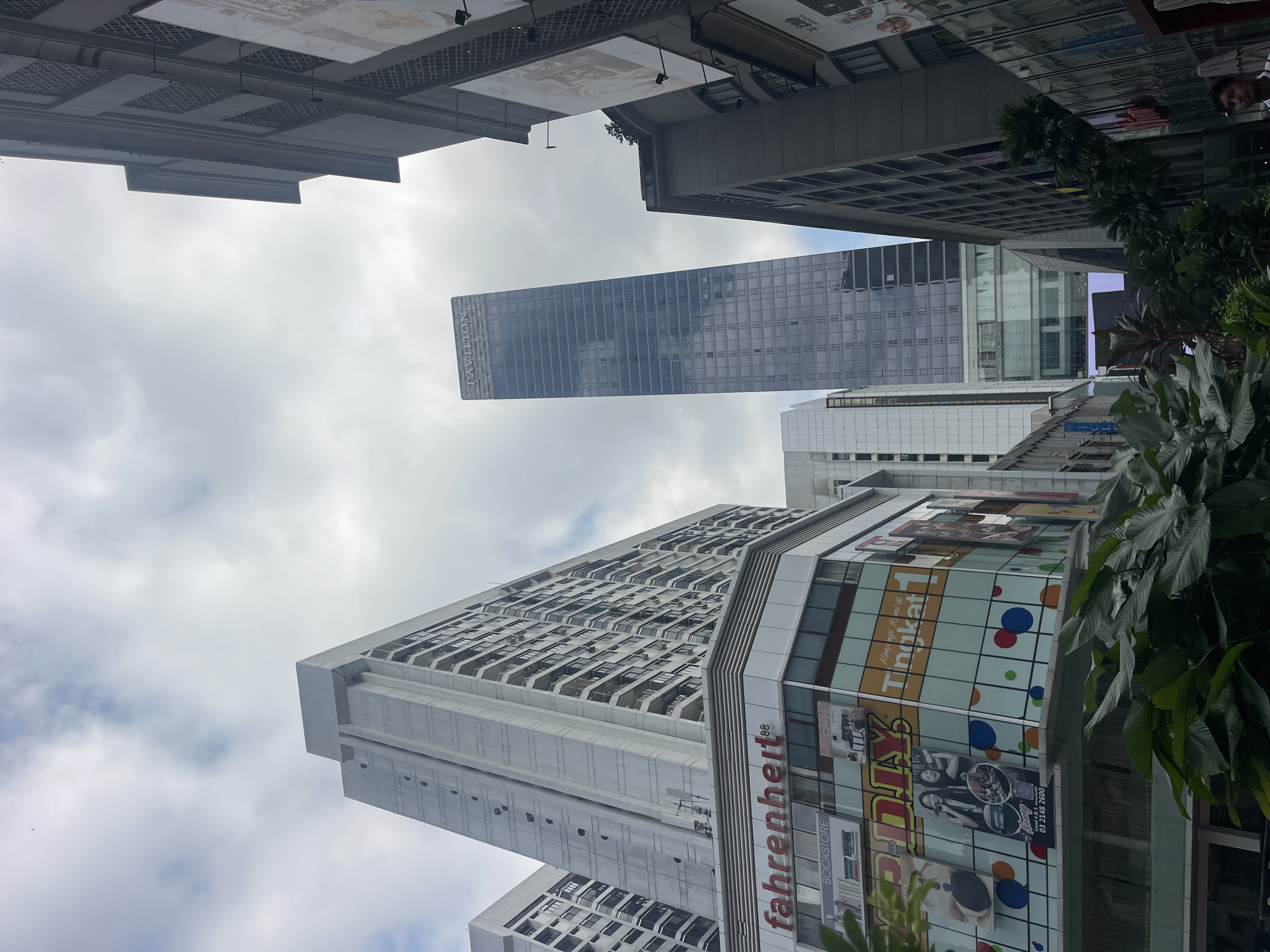 The image depicts a street-level view of a dense urban environment in Kuala Lumpur, Malaysia, dominated by towering skyscrapers and commercial buildings under a cloudy sky. In the foreground, lush green tropical foliage obscures the lower portions of several buildings.
The most prominent structures include a tall, slender, glass-facade skyscraper with "PAVILION" visible at its apex, suggesting it is part of the Pavilion Kuala Lumpur complex. To its left, a large, multi-story commercial building features a prominent sign for "fahrenheit 88" in red lettering, along with other visible signage for "BOOKSTORE," "CAFÉS," "MR. DIY," and "Tingkat 1." The facade of this building is adorned with colorful circles and advertisements featuring people and products.
To the far left, a very tall, concrete residential or office building with numerous balconies rises into the frame. To the right of the Pavilion skyscraper, another large, gray building with a grid-like facade is visible, and further to the right, part of a building with large advertising banners is seen.
The sky is overcast with scattered clouds. The street level appears to be a pedestrian area, with greenery suggesting a planned urban landscape design. A person in a white shirt is partially visible in the lower right corner, looking towards the right edge of the frame. The overall impression is one of a vibrant commercial and residential district in a major Asian city.