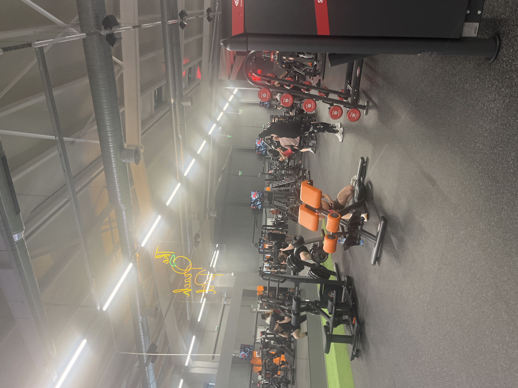 The image depicts the interior of a gym, likely located in Plovdiv, Bulgaria, given the context. The space is filled with various exercise machines, including weightlifting equipment, leg press machines, and potentially cardio machines. Several people are present, using the equipment. The gym features overhead lighting and ventilation ducts. A neon sign is also visible. The floor is covered with a dark, textured material.