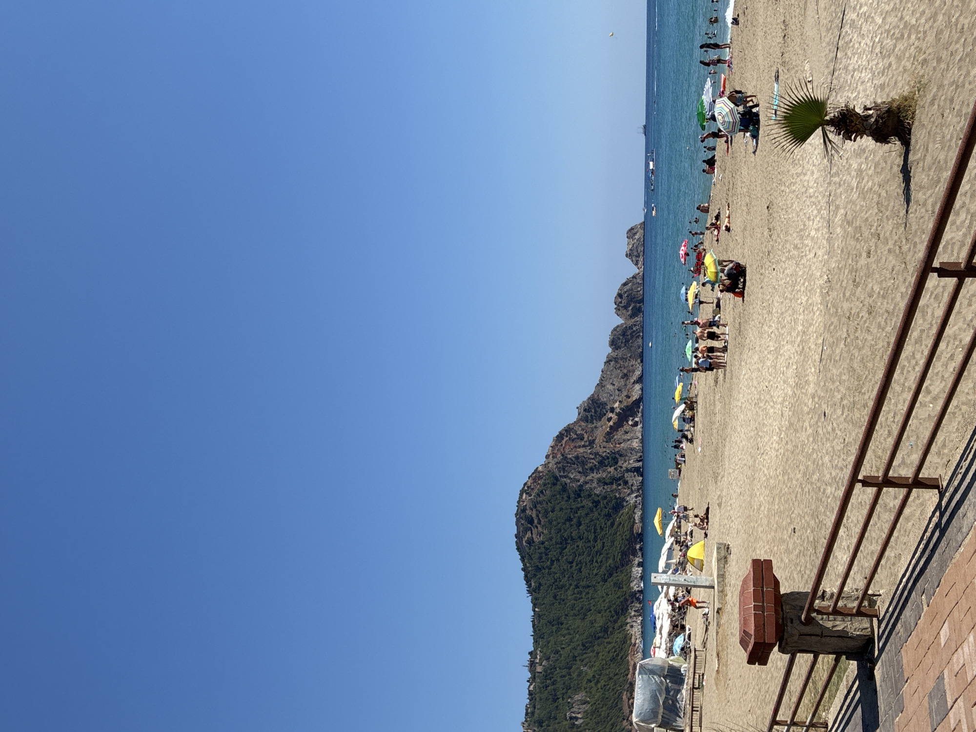 This image captures a sunny day at Cleopatra Beach in Alanya, Turkey. The foreground features a brown metal railing and a brick and stone pillar with a layered brick cap. Beyond this, a wide expanse of sand is populated with numerous beachgoers. People are scattered across the beach, some sitting on towels, others standing, and a few are visible in the shallow water. Beach umbrellas of various colors, including yellow, green, white, and patterned designs, provide shade. In the middle ground, the turquoise Mediterranean Sea is calm, with a few small boats visible in the distance. Dominating the background is a large, verdant hill or small mountain, covered in dense green trees, with rocky outcrops and the suggestion of ancient structures on its summit. The sky is a clear, vibrant blue.