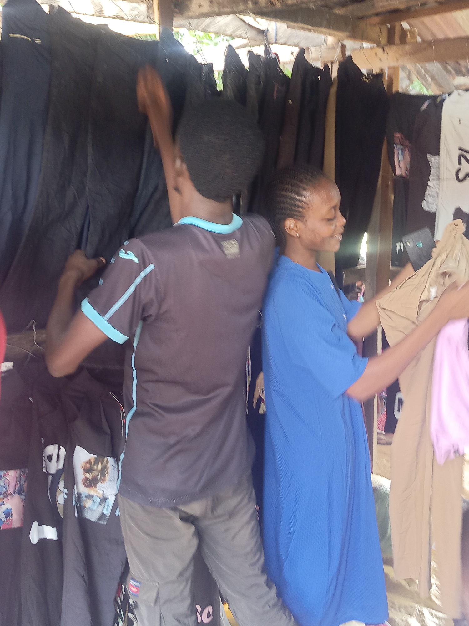 Two people, a male and female, stand close together in what appears to be a small outdoor clothing shop. The man is on the left, with his back to the camera. He wears a dark grey short-sleeved shirt with light blue trim and grey pants. He is reaching towards a rack of dark-colored clothing. The woman, on the right, faces right and smiles. She wears a blue dress and holds tan and pink colored pieces of clothing. Behind them are clothes hanging on racks. The setting is a simple wooden structure. The location is Akwanga, Nigeria.