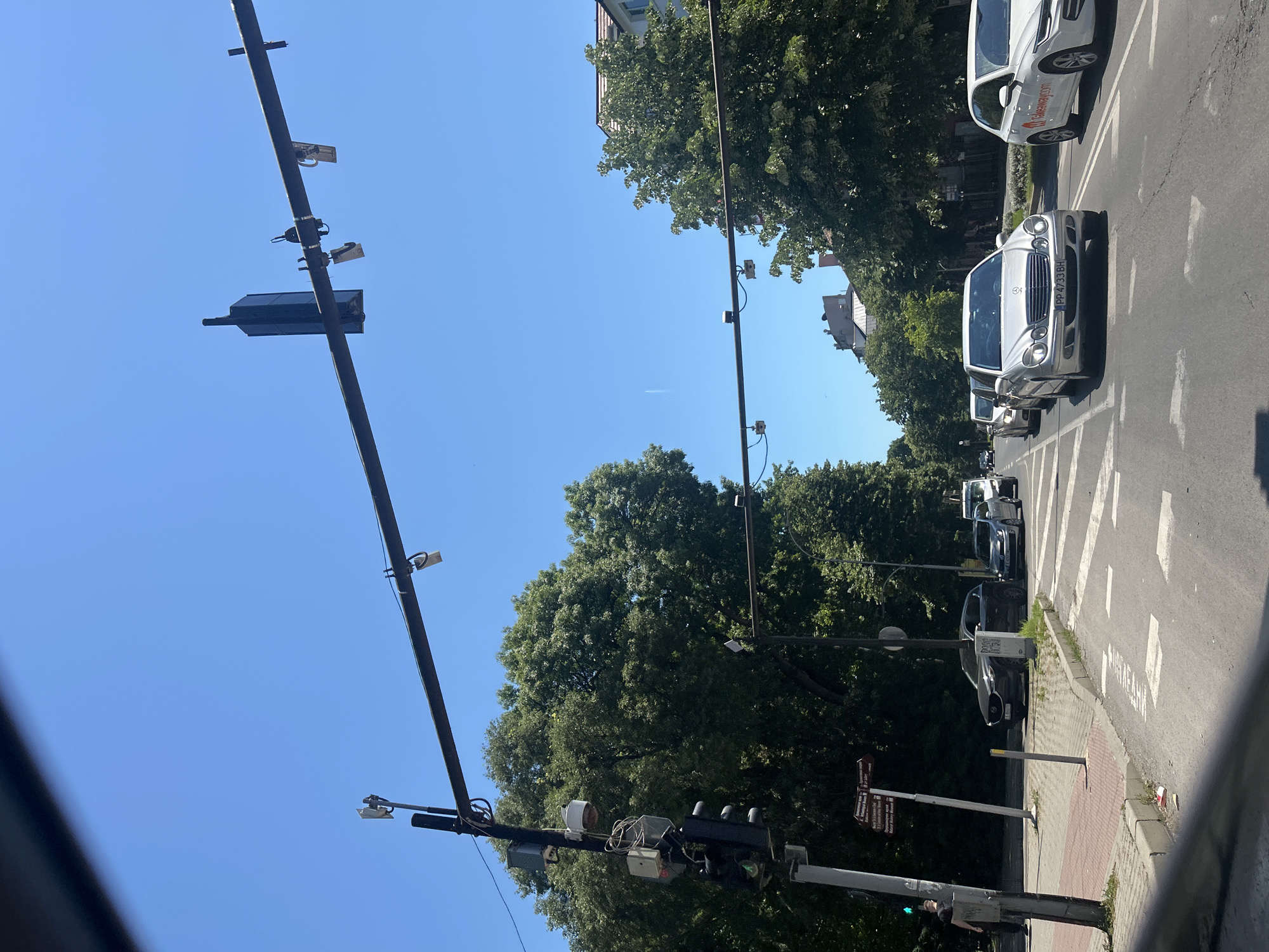 The image depicts a street scene in Varna, Bulgaria, under a clear blue sky. A traffic light pole with several attached devices, including cameras and what appears to be a siren, stands on the left side of the road. Overhead, a black horizontal bar stretches across the street, fitted with traffic cameras or sensors. Several cars are visible on the road, moving in the same direction. The street is lined with trees and some buildings are visible in the background. There is a sidewalk on the left, with a person standing on it. The road has pedestrian crossing markings and traffic lanes.